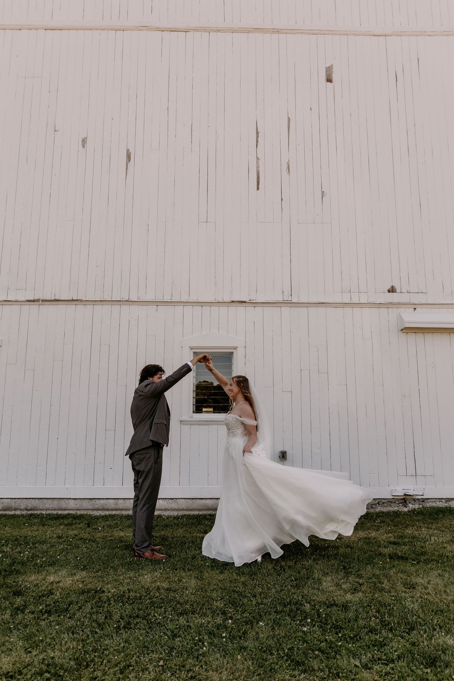 Bride and groom dancing outdoors in front of a white barn, bride's gown flowing in the breeze.
