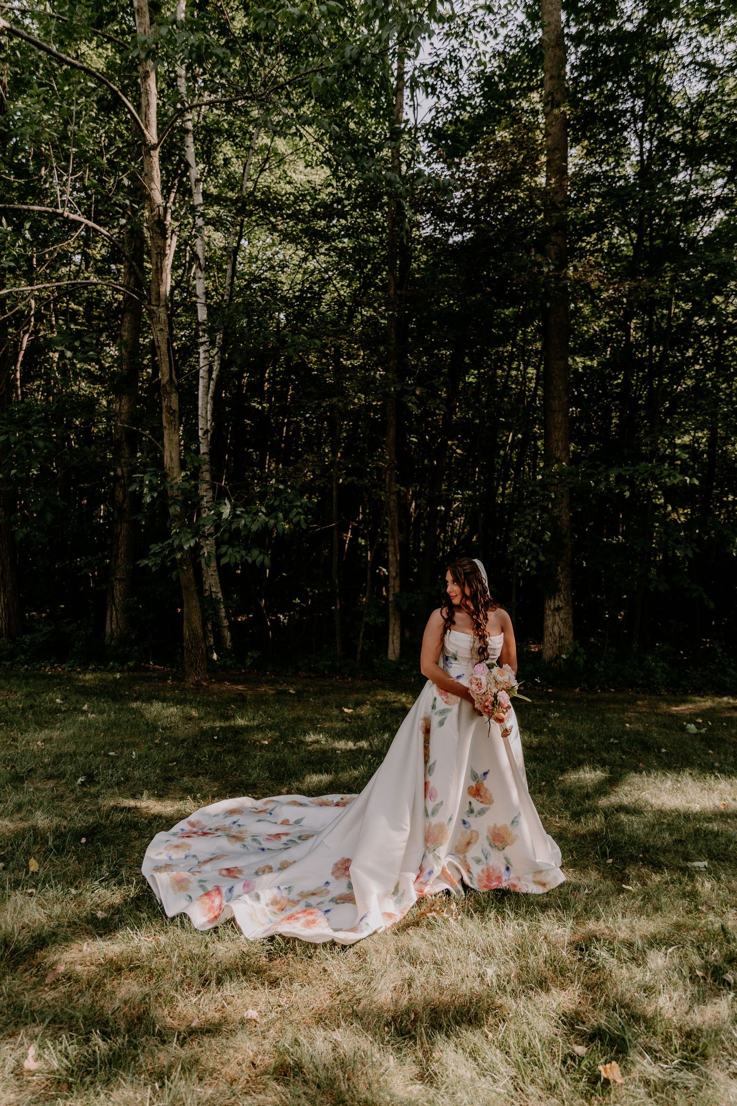 Bride in a stunning floral wedding gown with a long train standing in a sunlit forest clearing.