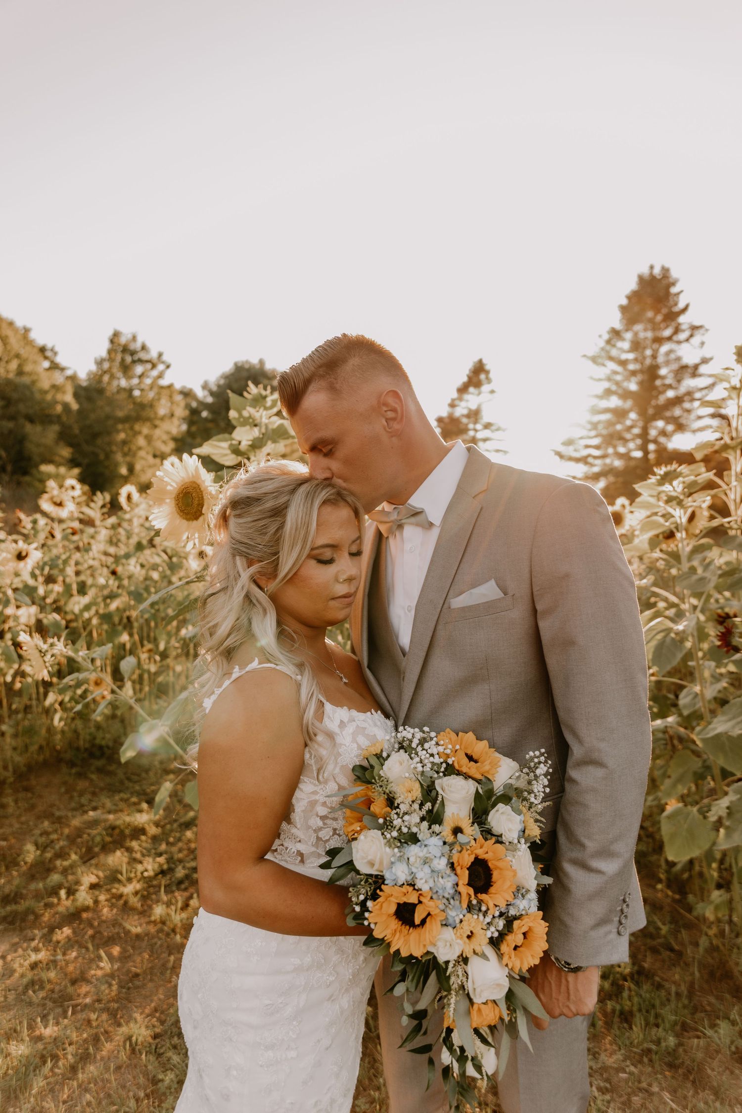 Bride and groom embrace in a sunflower field, she holds a bouquet of sunflowers and white roses in golden hour light.