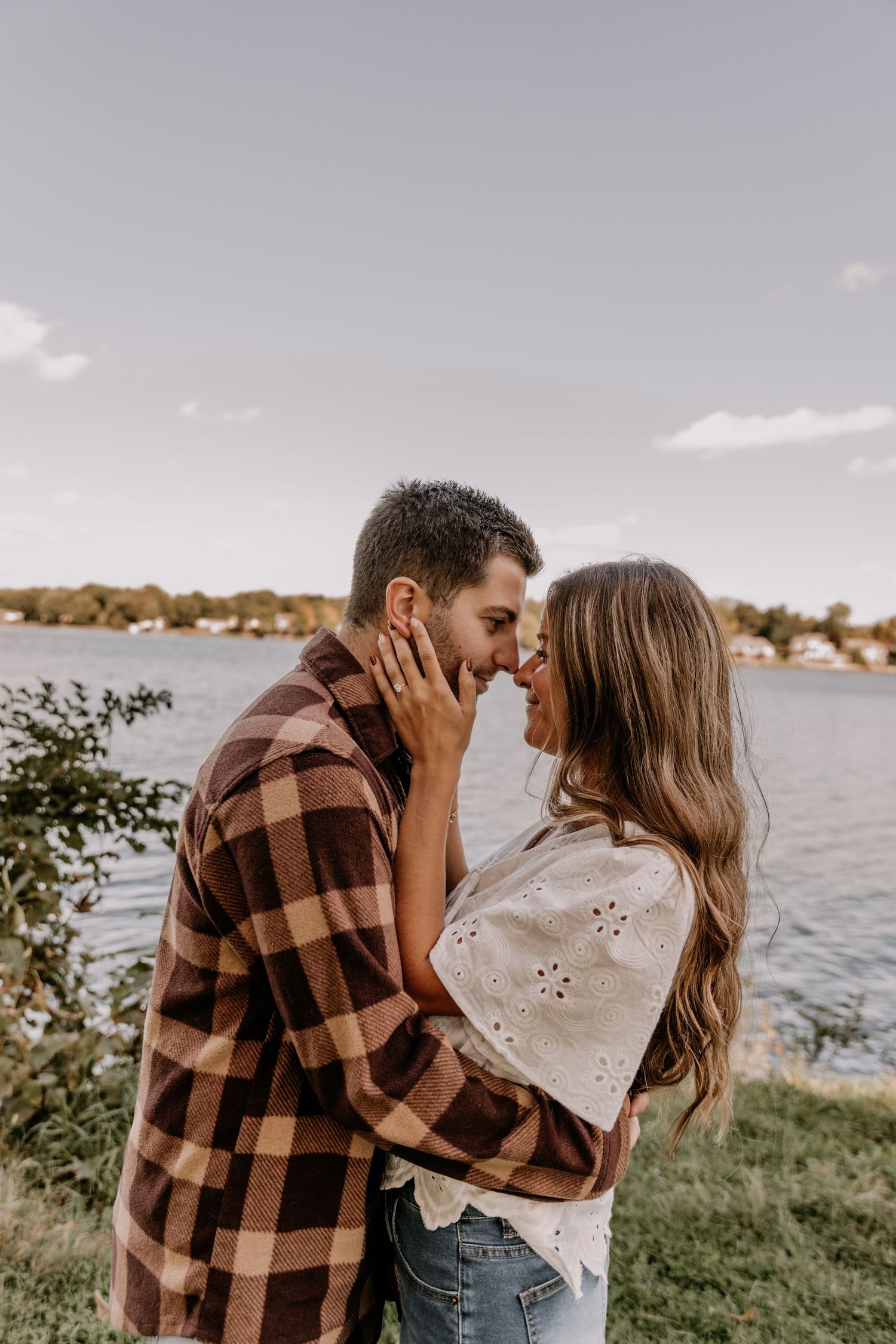 Couple sharing a romantic moment by a lakeside, man in plaid shirt and woman in white lace top touching foreheads.