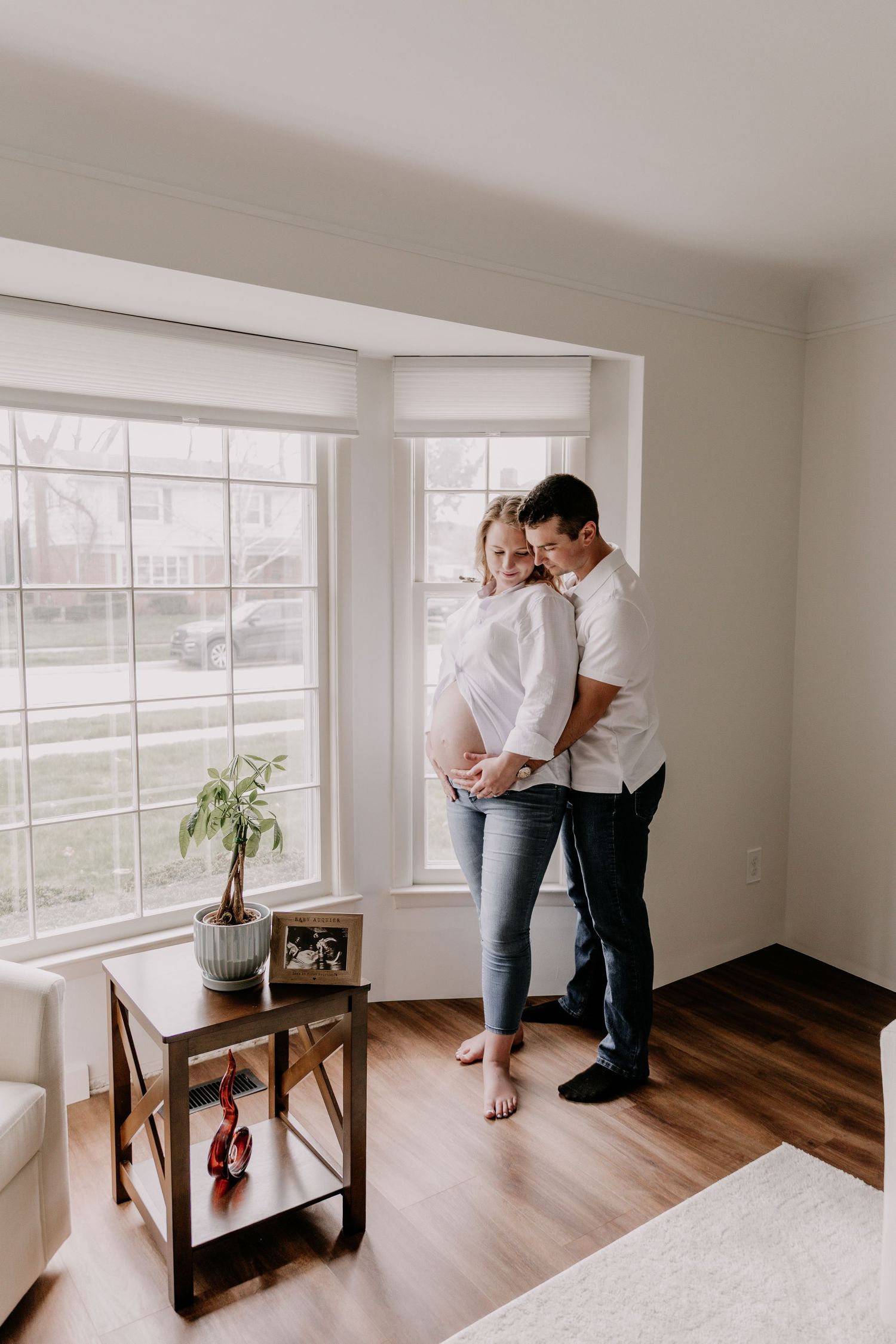 Pregnant couple embracing by large window in modern living room with hardwood floors and natural light.