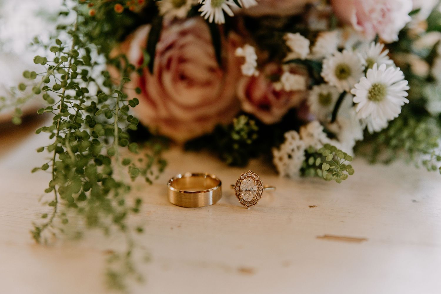 Gold wedding bands and floral bouquet with pink roses and white daisies on a wooden surface.