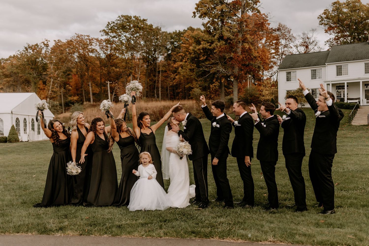 Wedding party in black attire poses together on grassy field during autumn with white building in background.