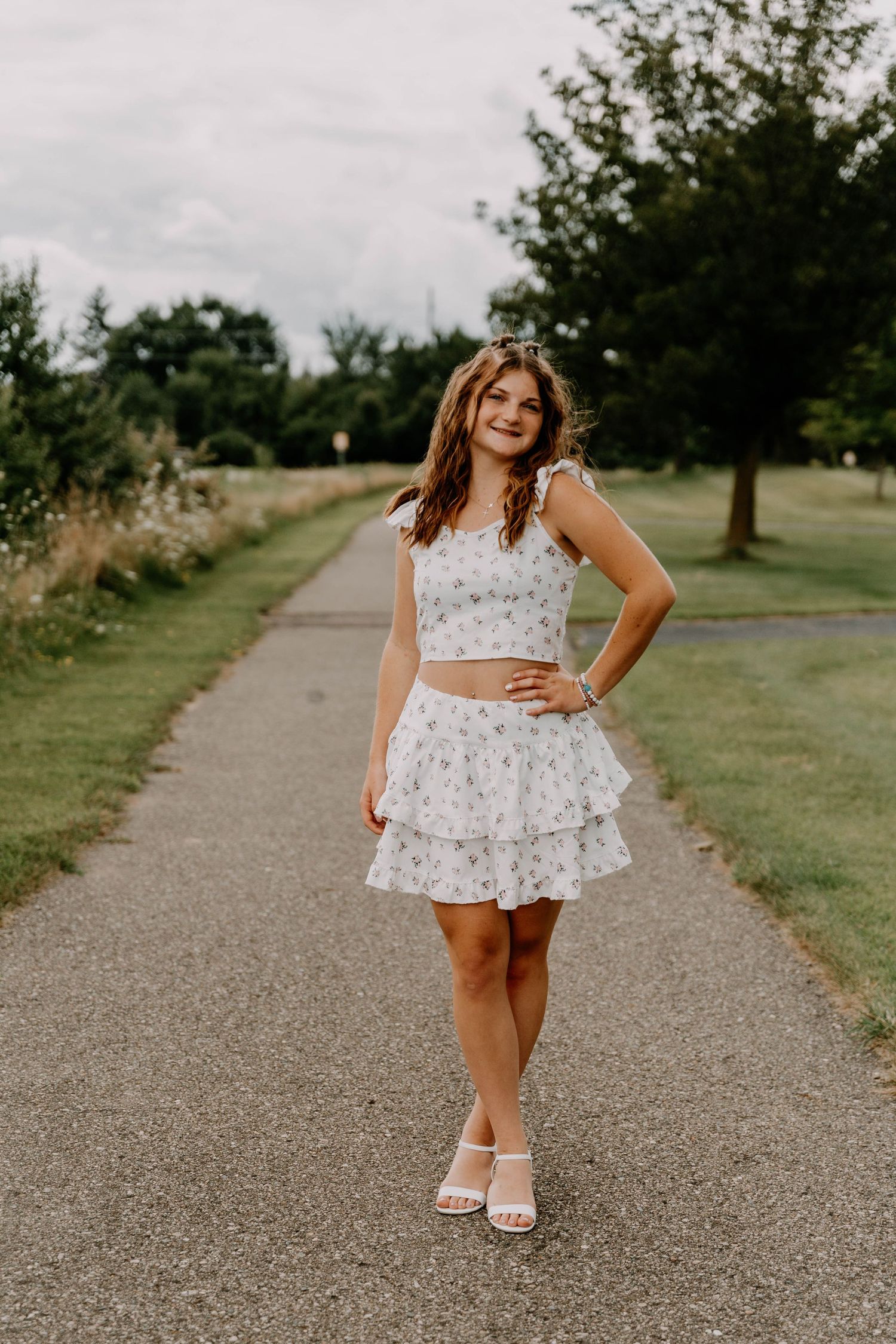 A person wearing a white floral dress poses on a gravel path surrounded by lush greenery.