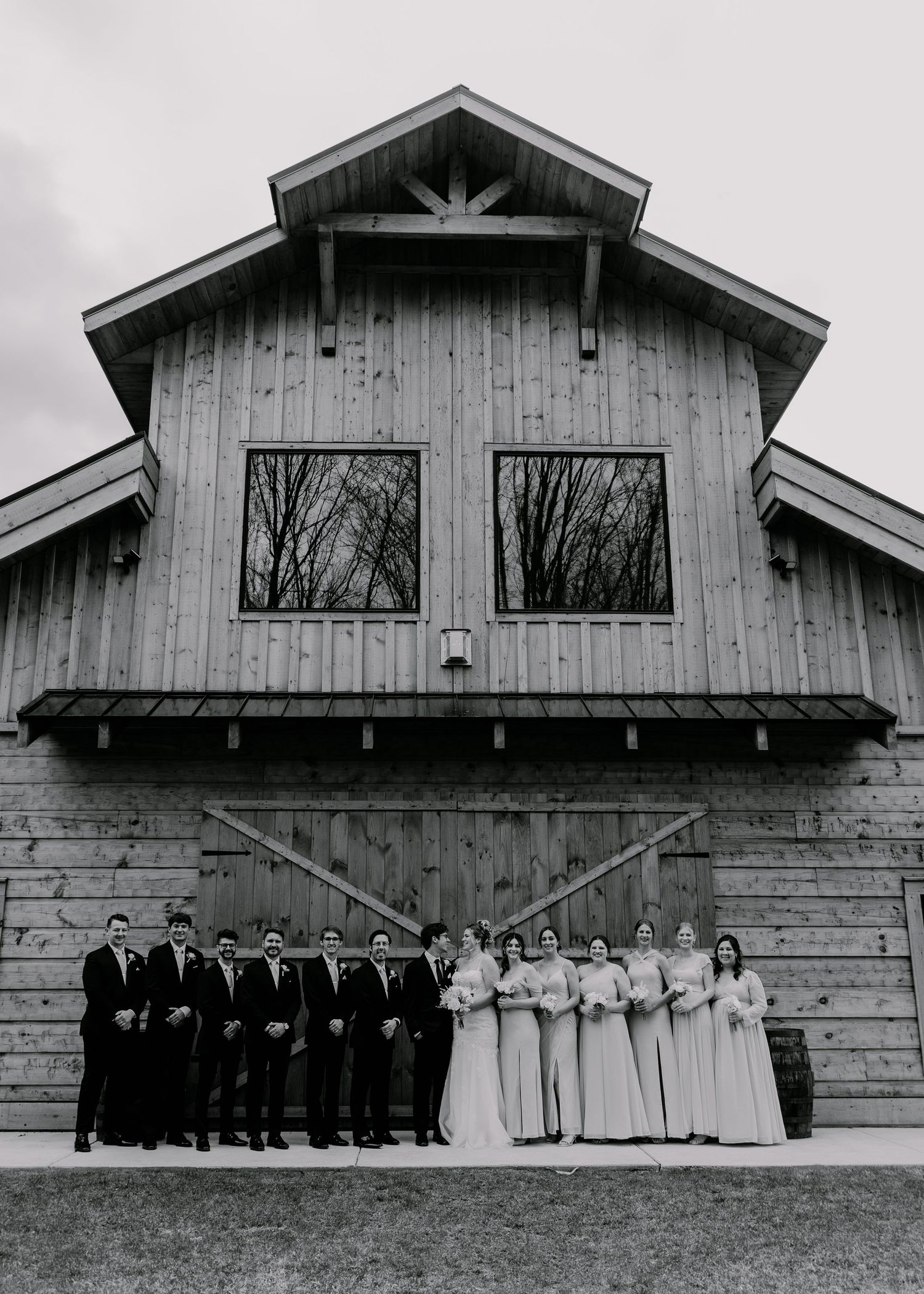 Wedding party poses in formal attire outside rustic barn in black and white photography.