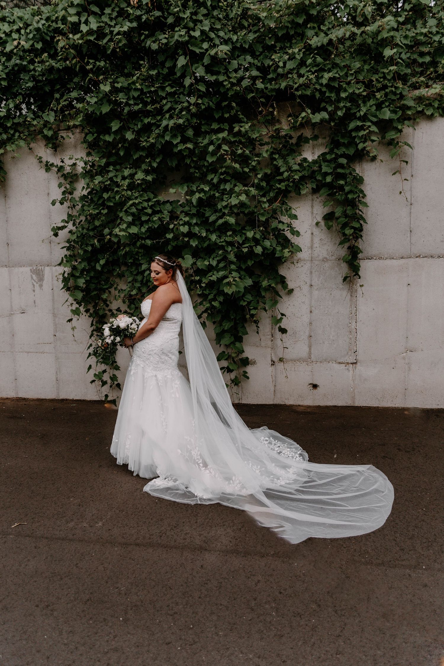 Elegant bride in flowing white gown poses against ivy-covered wall.