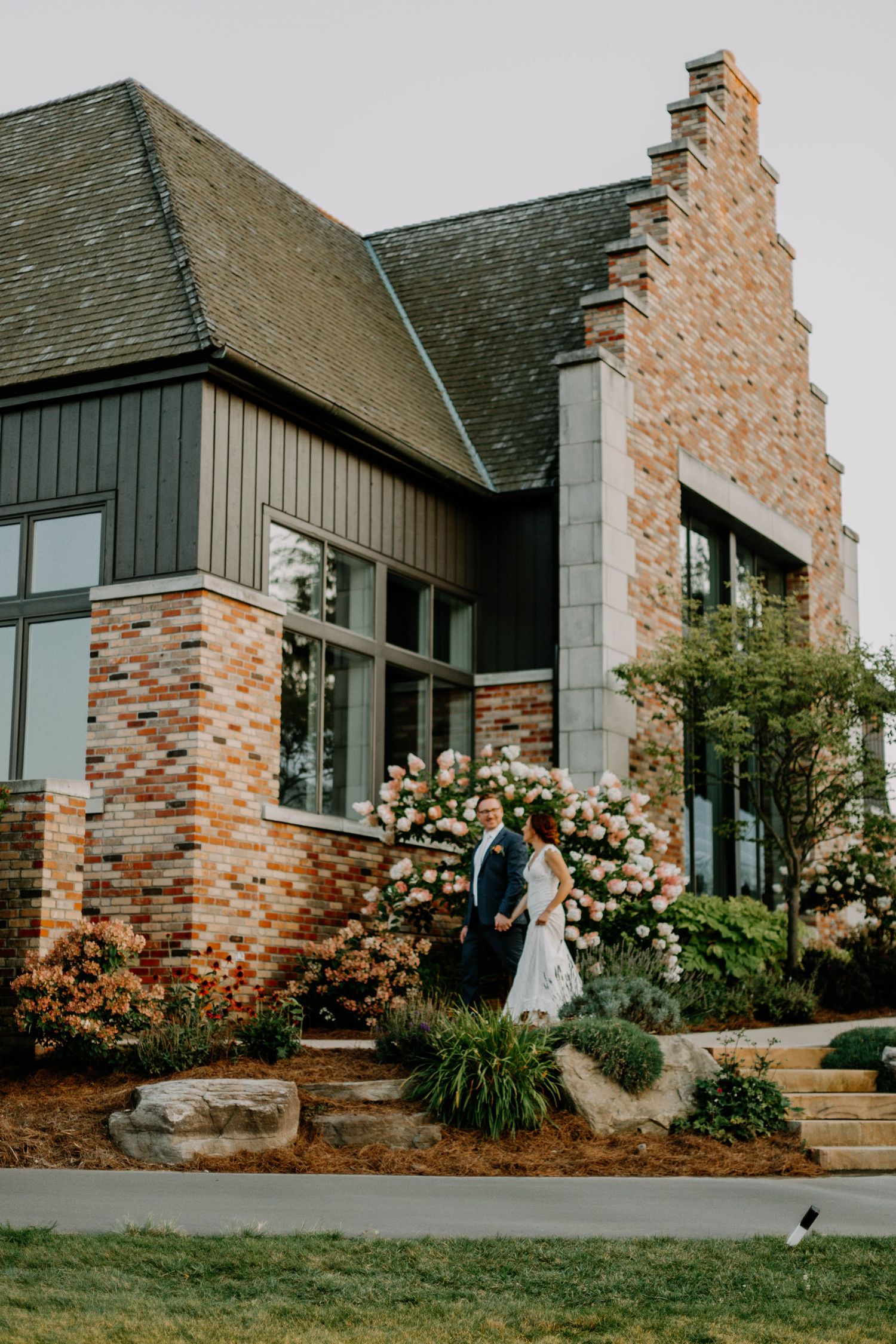 A bride and groom pose outside a modern stone and brick venue with climbing roses.