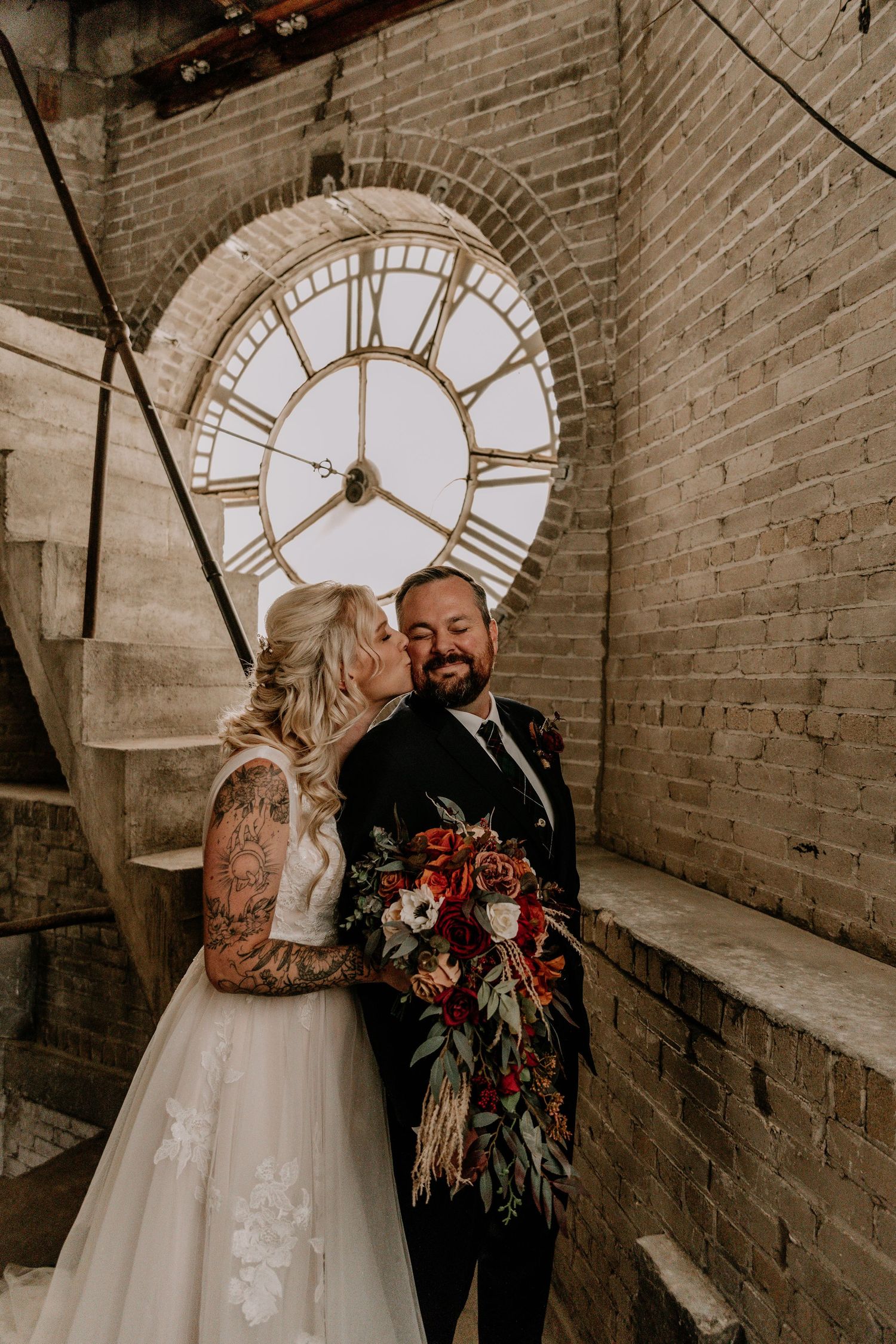 A couple shares an intimate moment in front of a vintage clock face in a brick-walled setting.