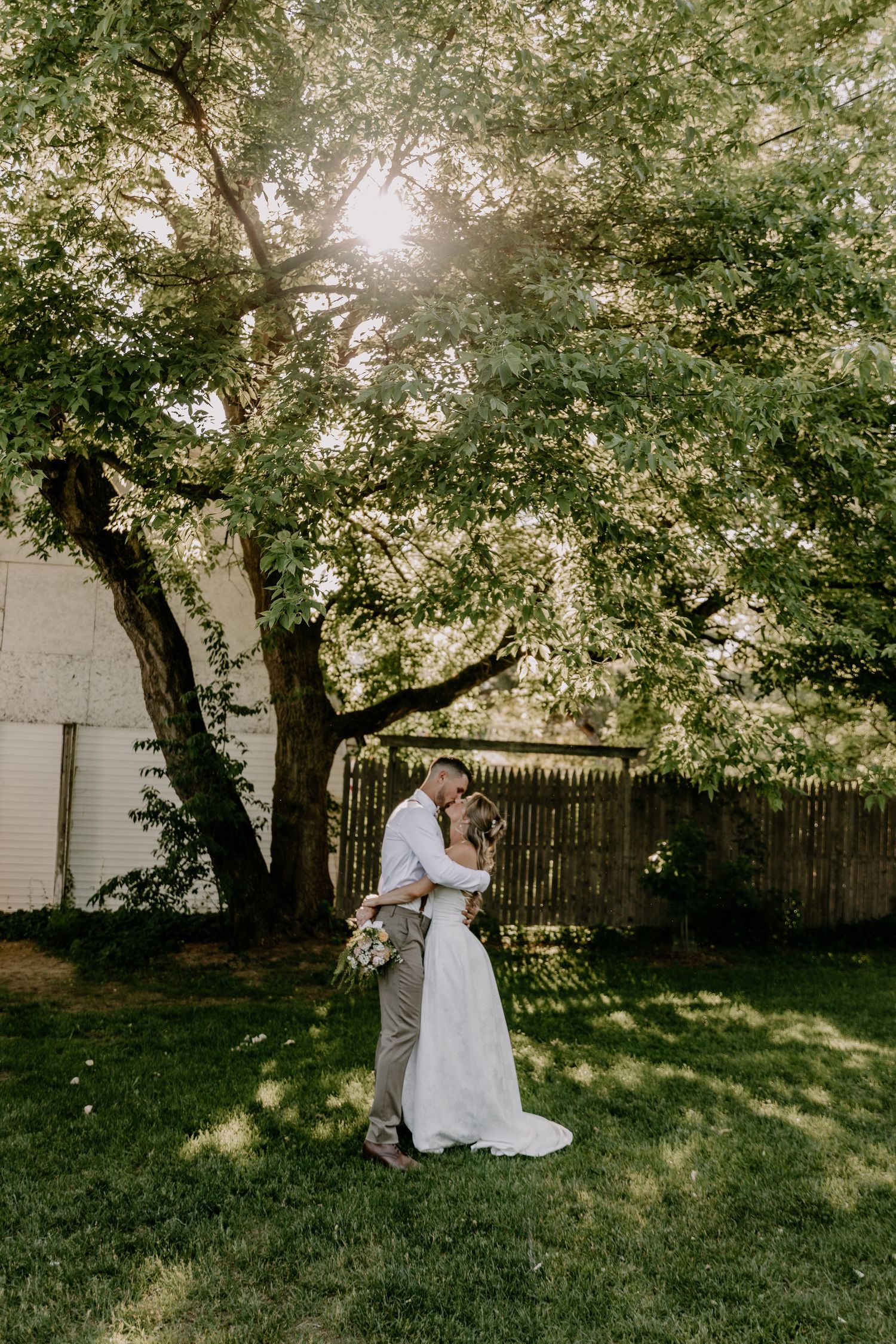 Bride and groom embracing under a large sunlit tree in a lush green backyard on their wedding day.