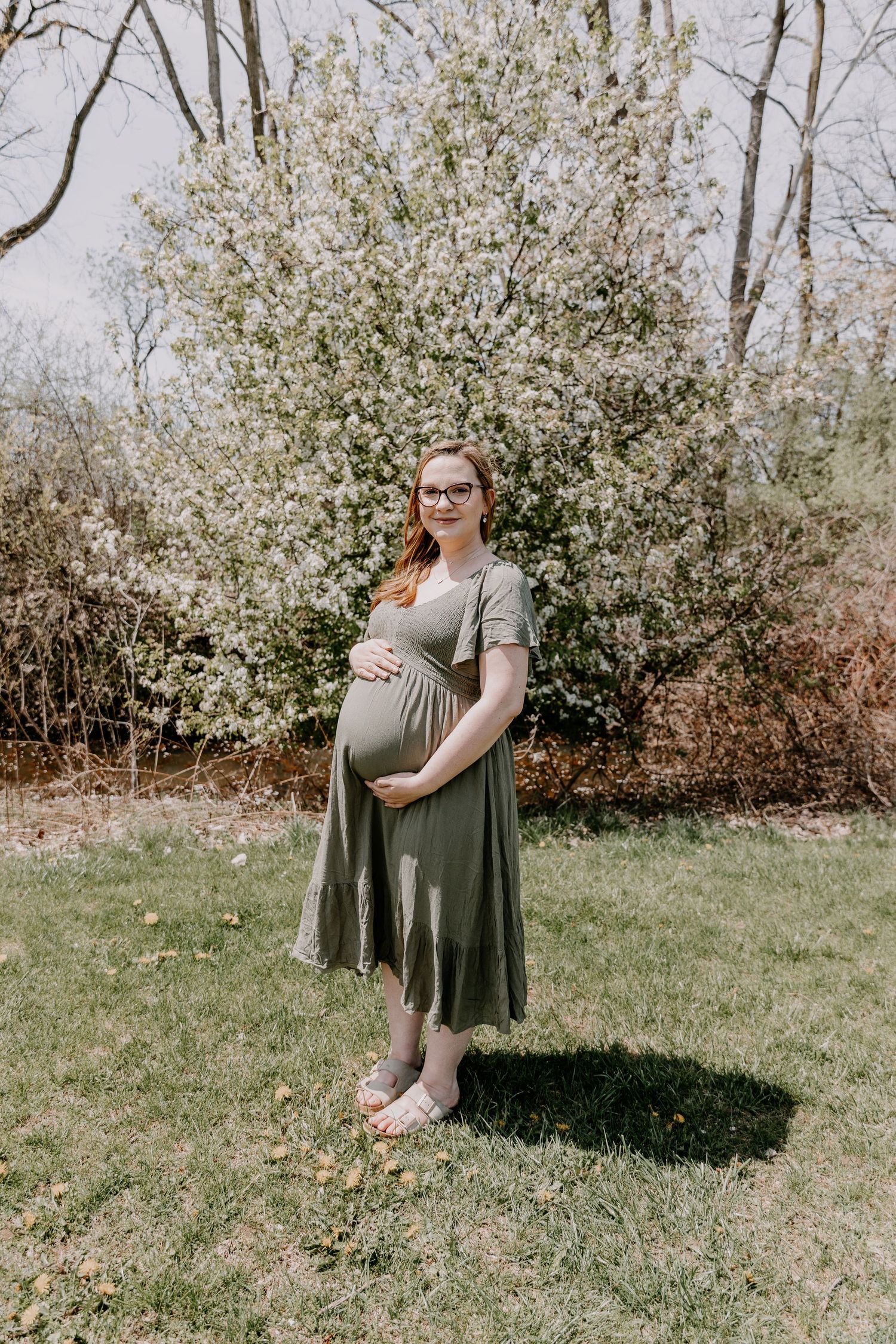 Pregnant woman in olive green dress cradling baby bump outdoors near blooming white tree on green lawn in spring.