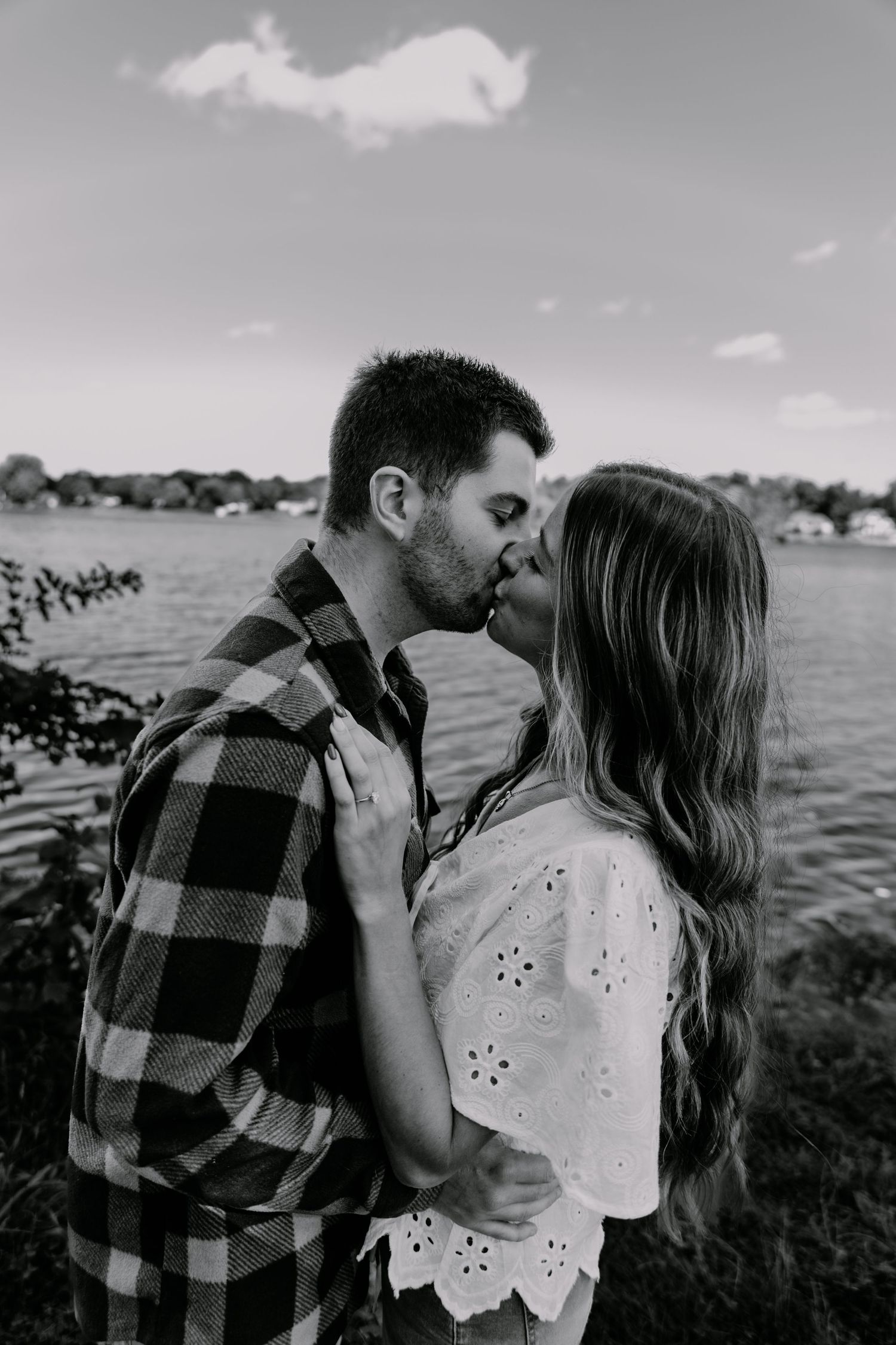 Black and white photo of a couple kissing near a lake, man in plaid shirt and woman in white lace dress.