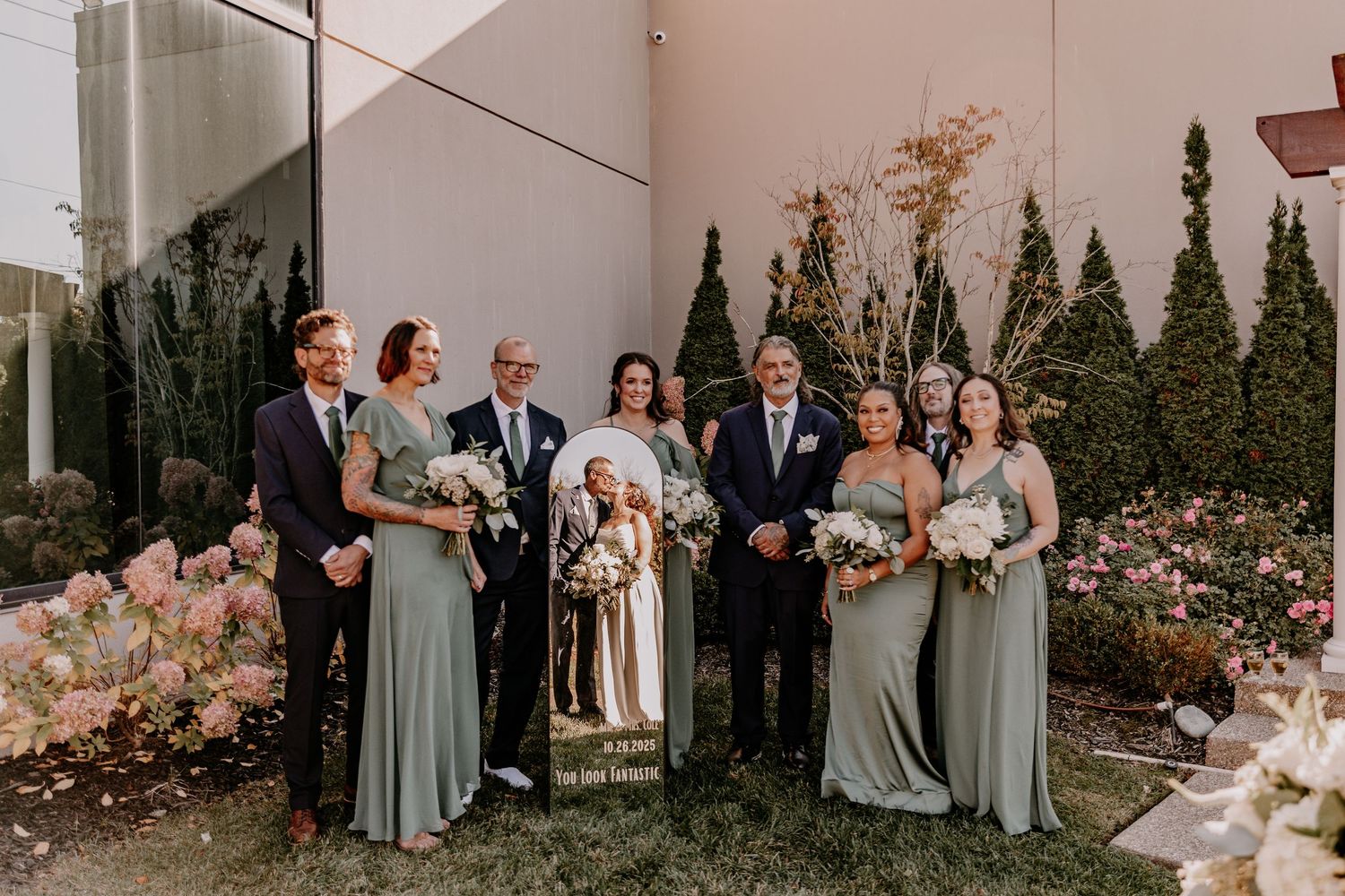 Wedding party members in sage green dresses and dark suits pose together in an outdoor garden setting.