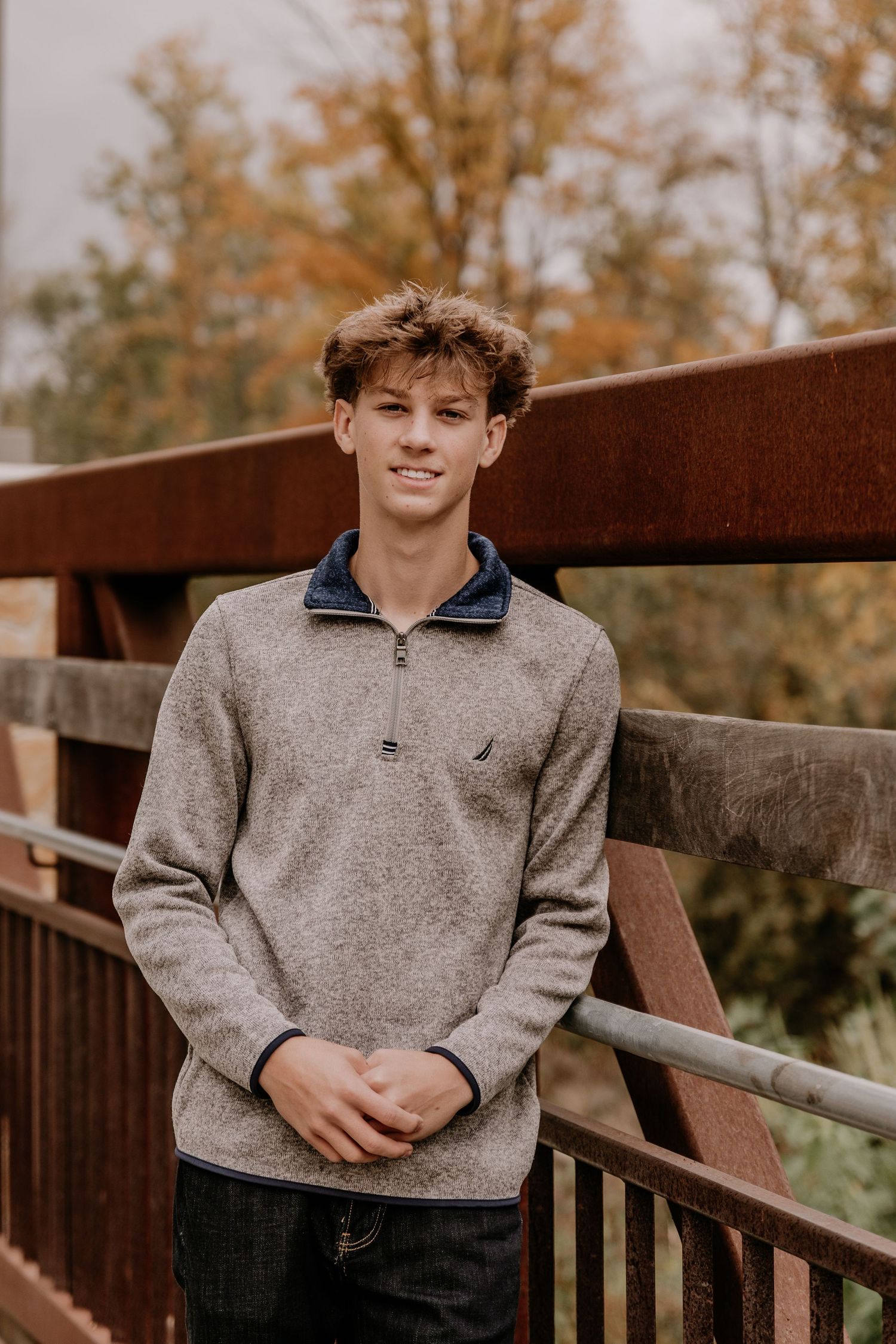 A person in a grey sweater sits on a wooden deck railing surrounded by autumn trees in the background.