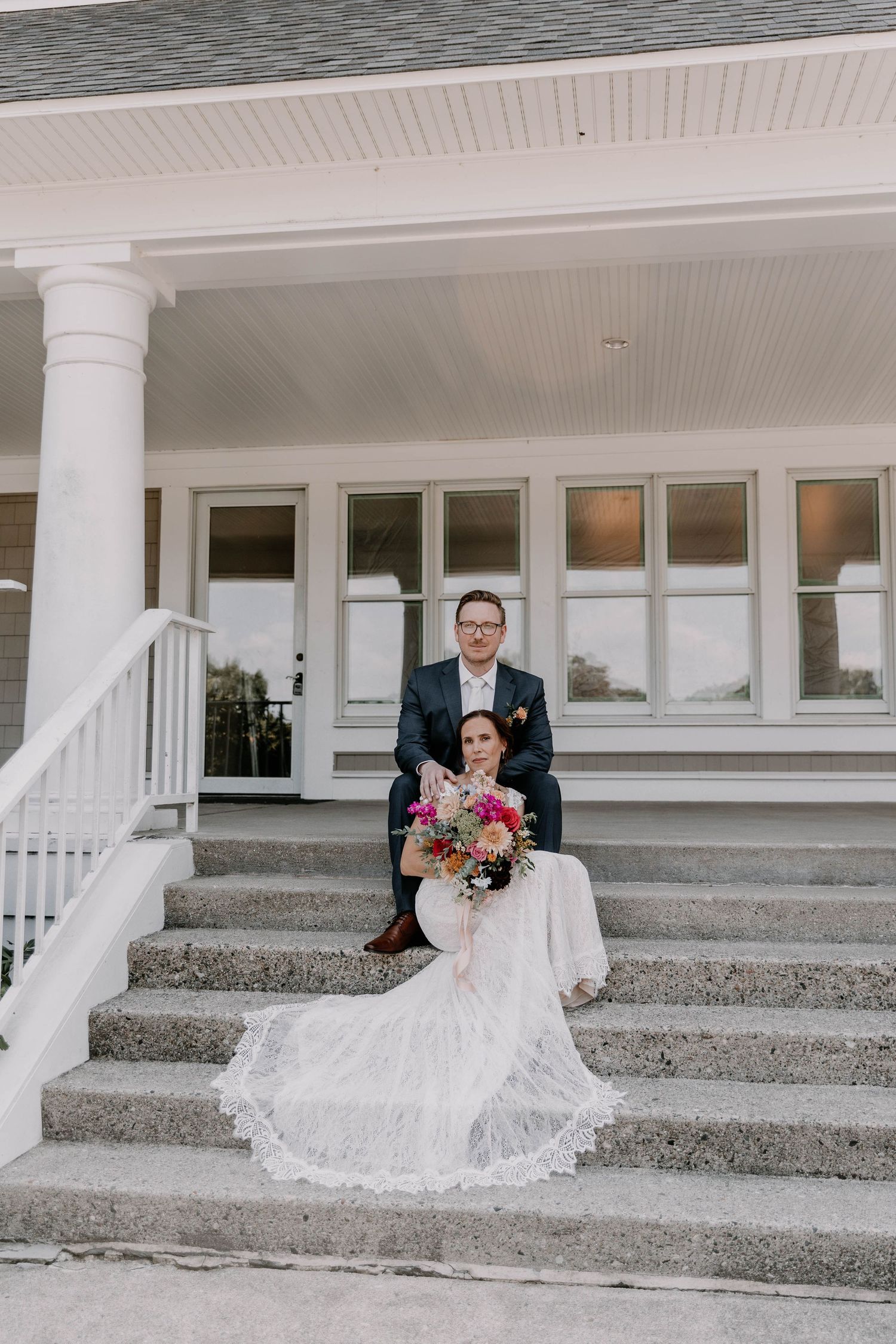 Newlyweds pose on white steps of an elegant venue with columns and a flowing bridal veil.