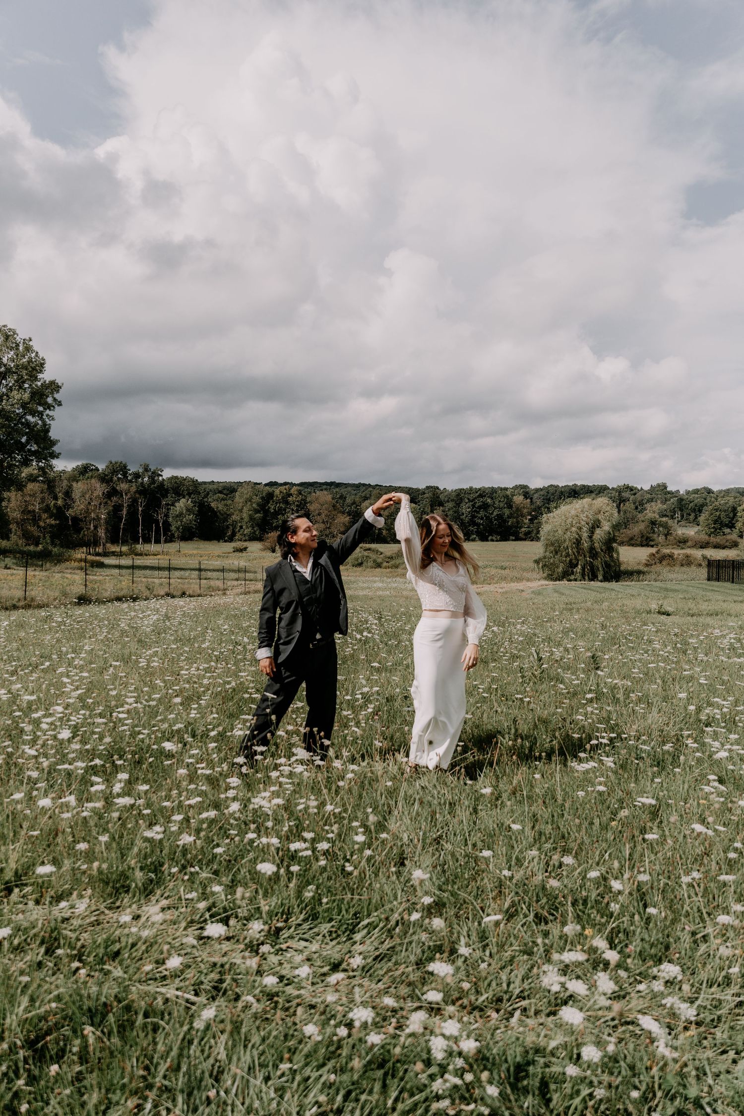 Couple dancing in wildflower meadow under dramatic cloudy sky.