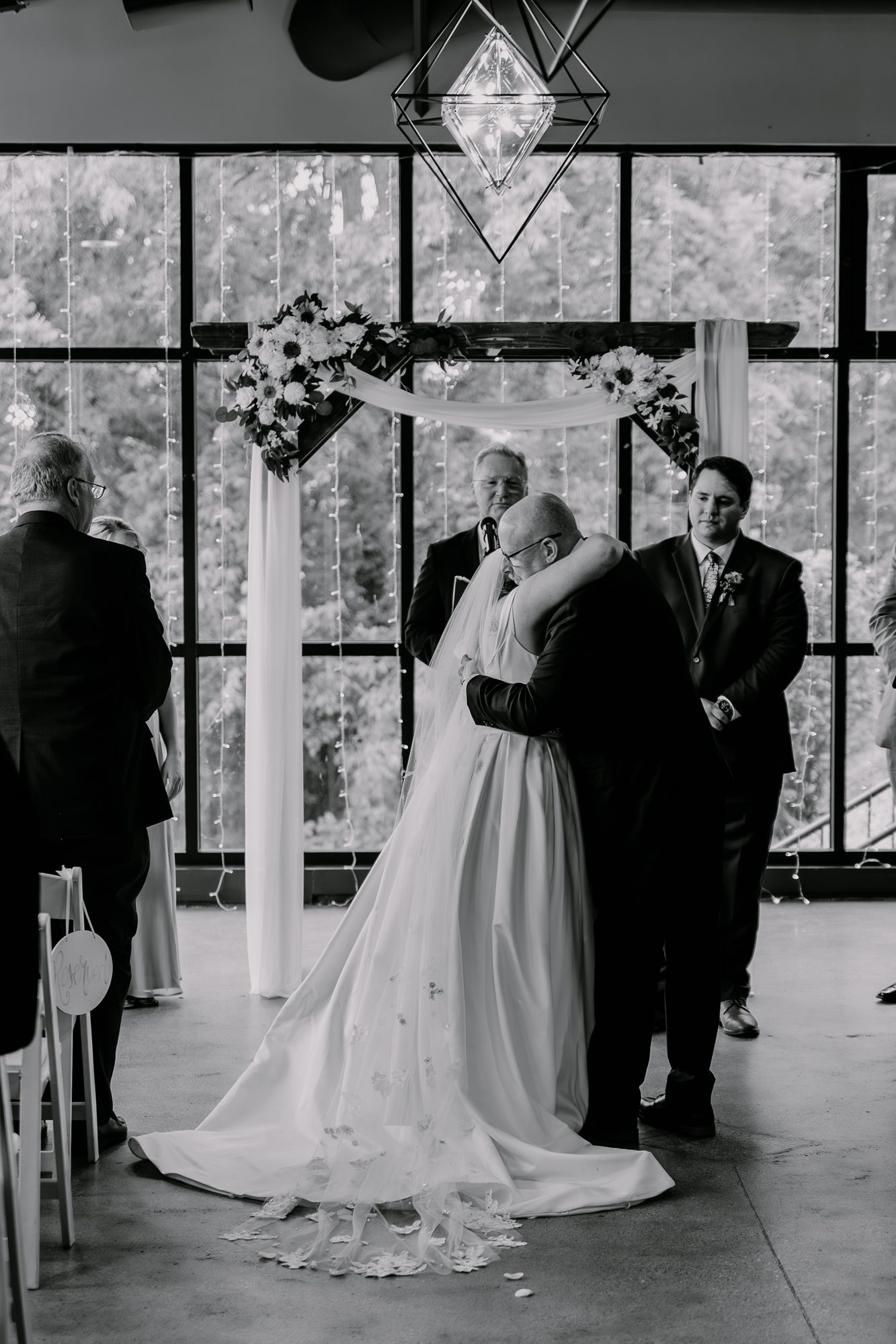 Wedding ceremony in a sunlit venue with floor-to-ceiling windows and geometric hanging lights.