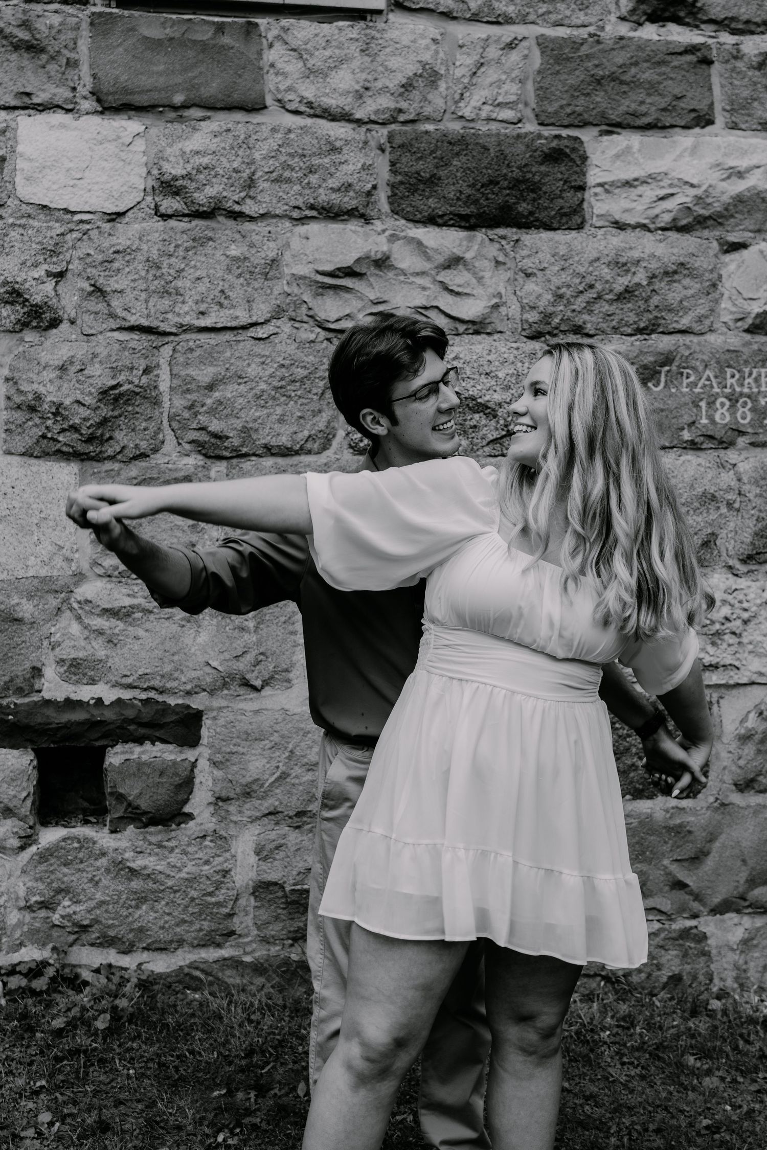 A couple strikes a playful dancing pose against a stone wall in black and white.