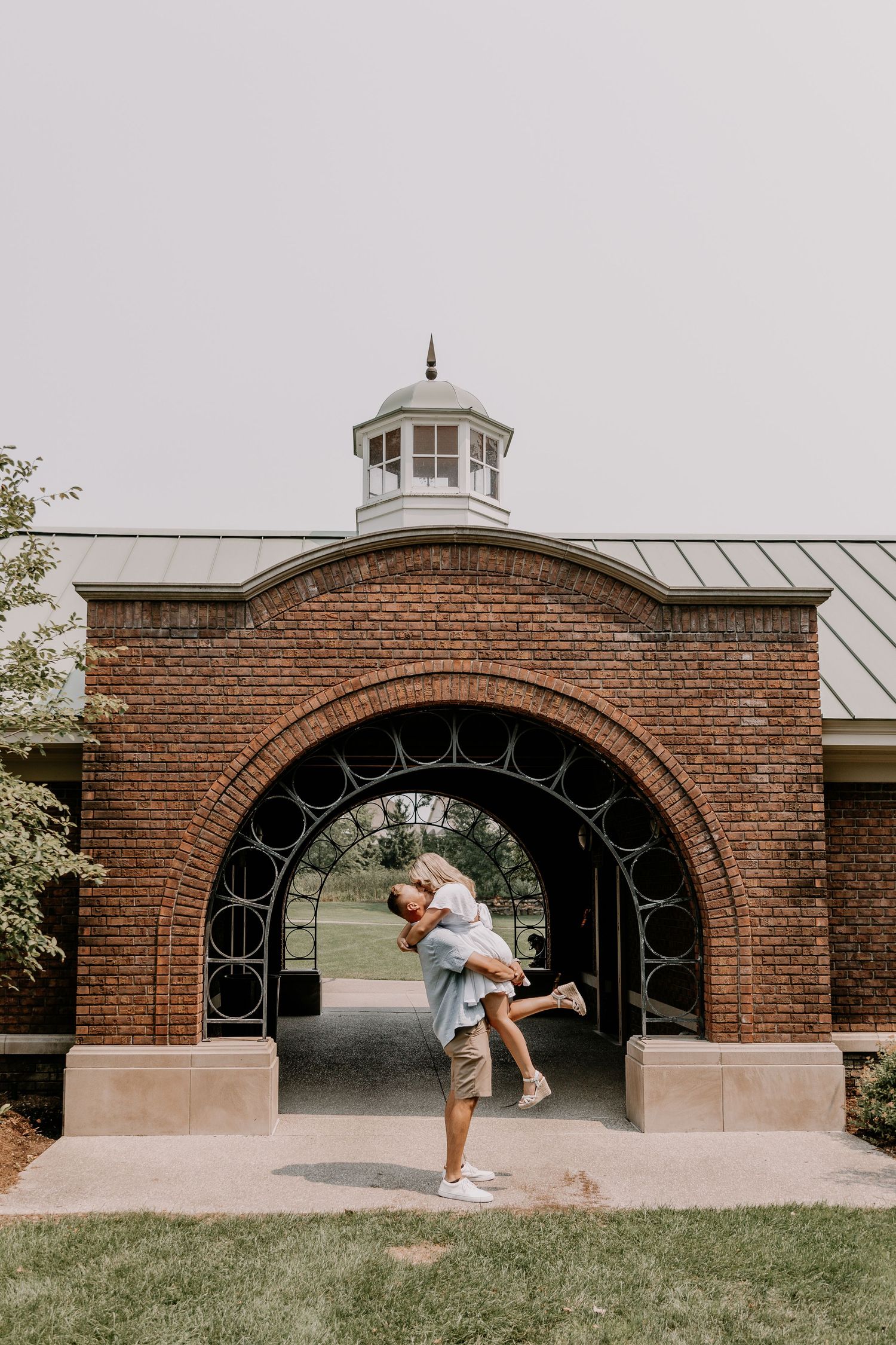 A couple embraces romantically under a brick archway with a white cupola on top at an elegant outdoor venue.