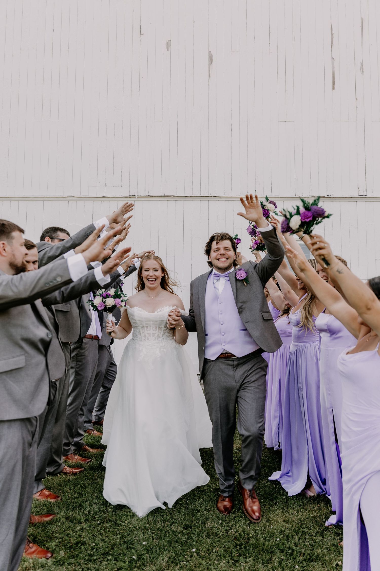 Bride and groom walk through a tunnel of raised hands with wedding party in gray suits and lavender dresses outdoors.