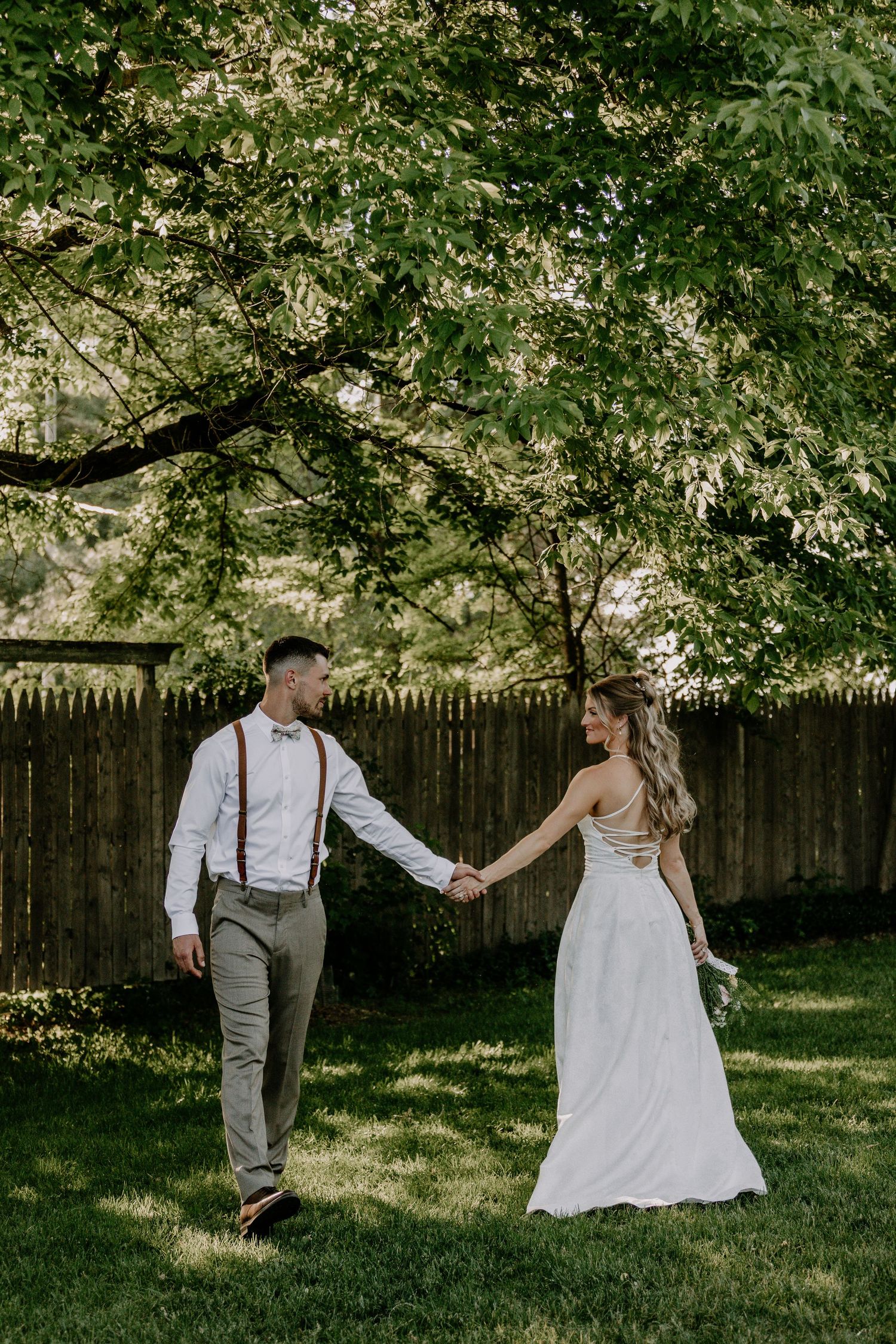 Bride and groom hold hands walking through lush green garden, bride in backless white gown, groom in suspenders.