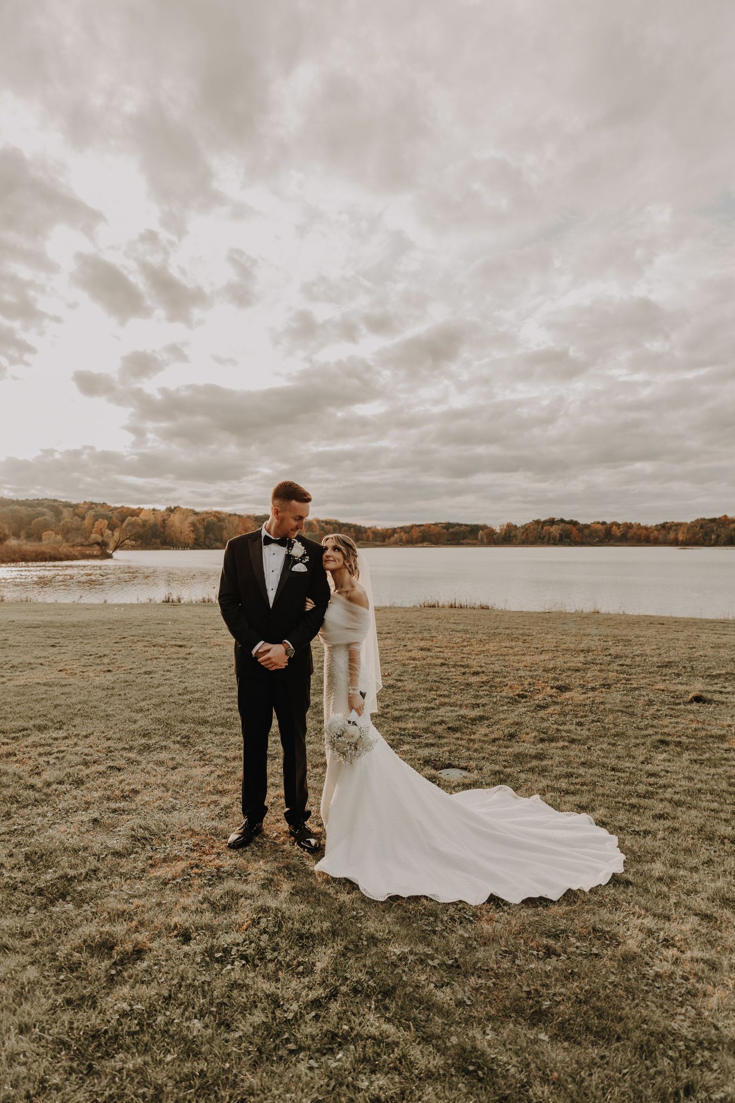 Bride in white gown with train and groom in black tuxedo pose by a lakeside on a cloudy autumn day.