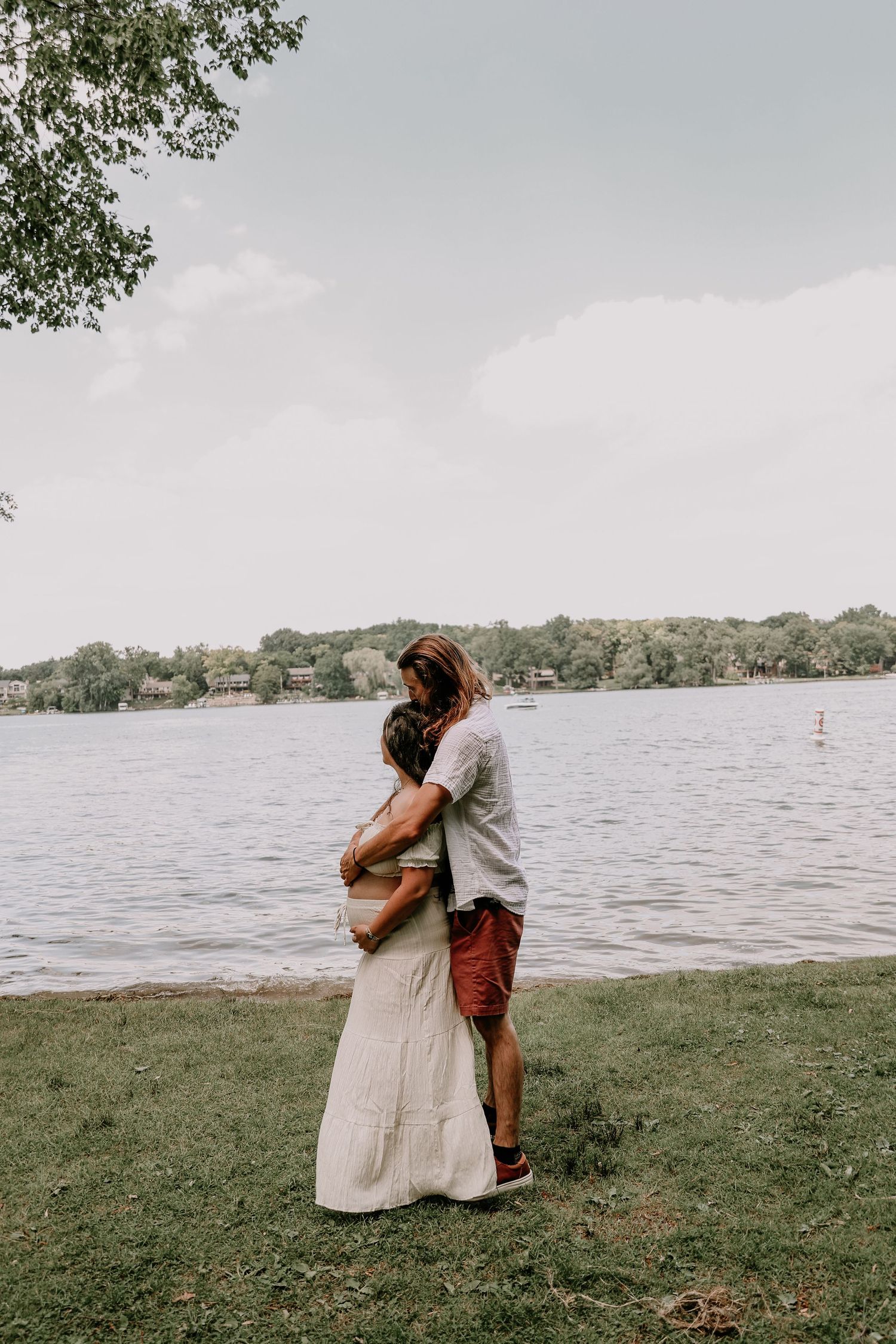 Couple embracing by a serene lake shore, surrounded by lush green trees on a cloudy summer day.