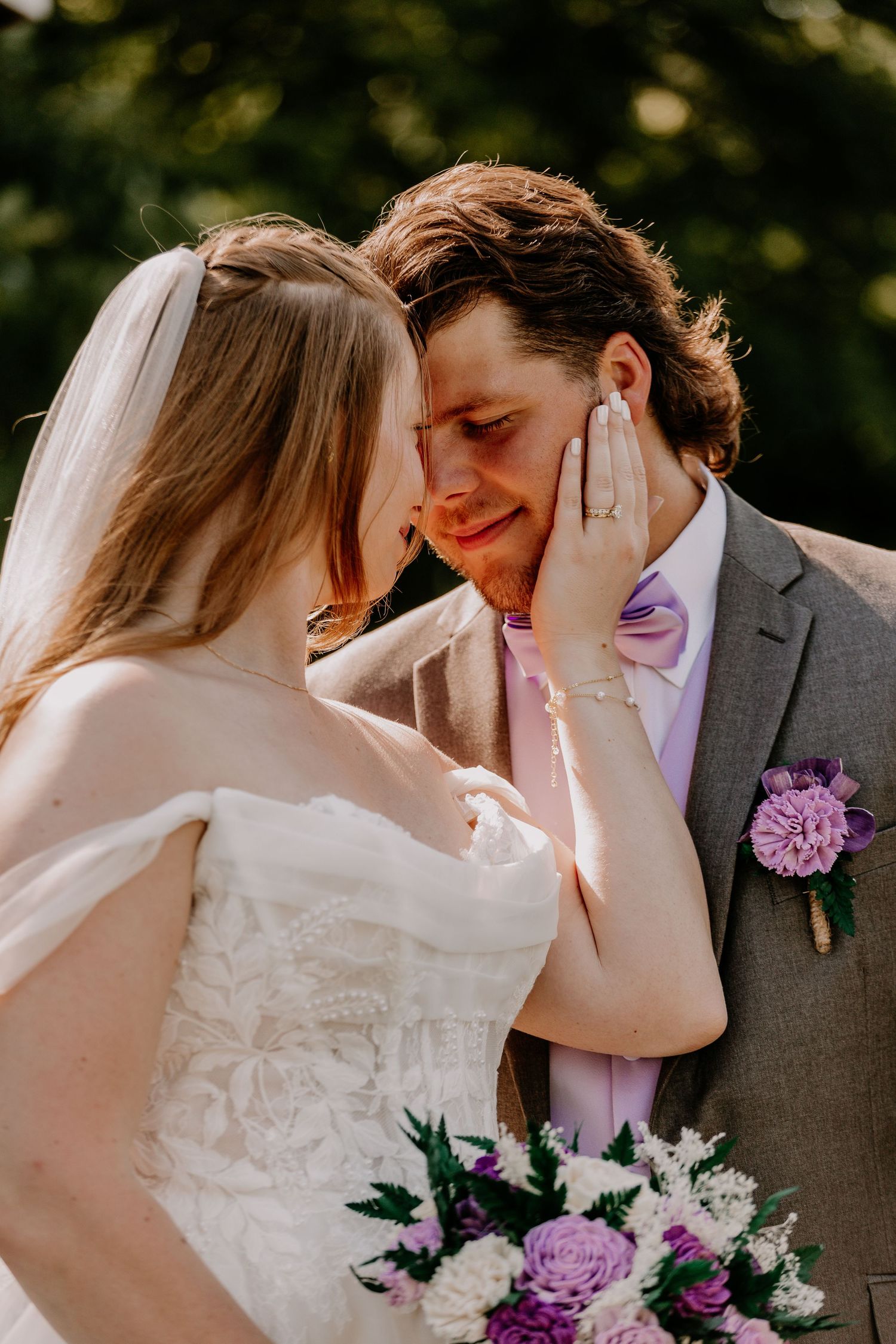 Bride tenderly cups grooms face as they share an intimate moment, with purple wedding bouquet and boutonniere.