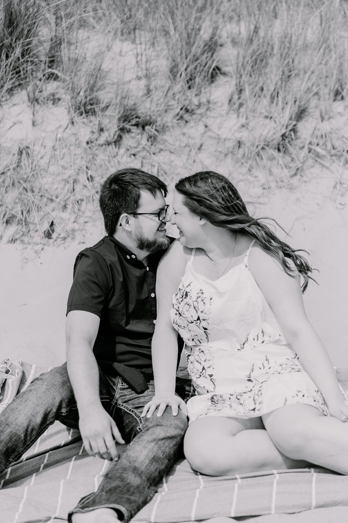 A black and white photo of a couple sitting close together on a blanket among beach grass.