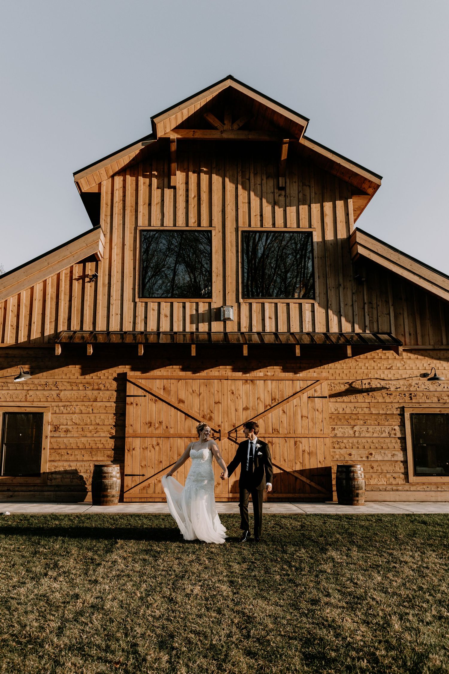 A bride and groom pose outside a warm-toned wooden barn with stone foundation.