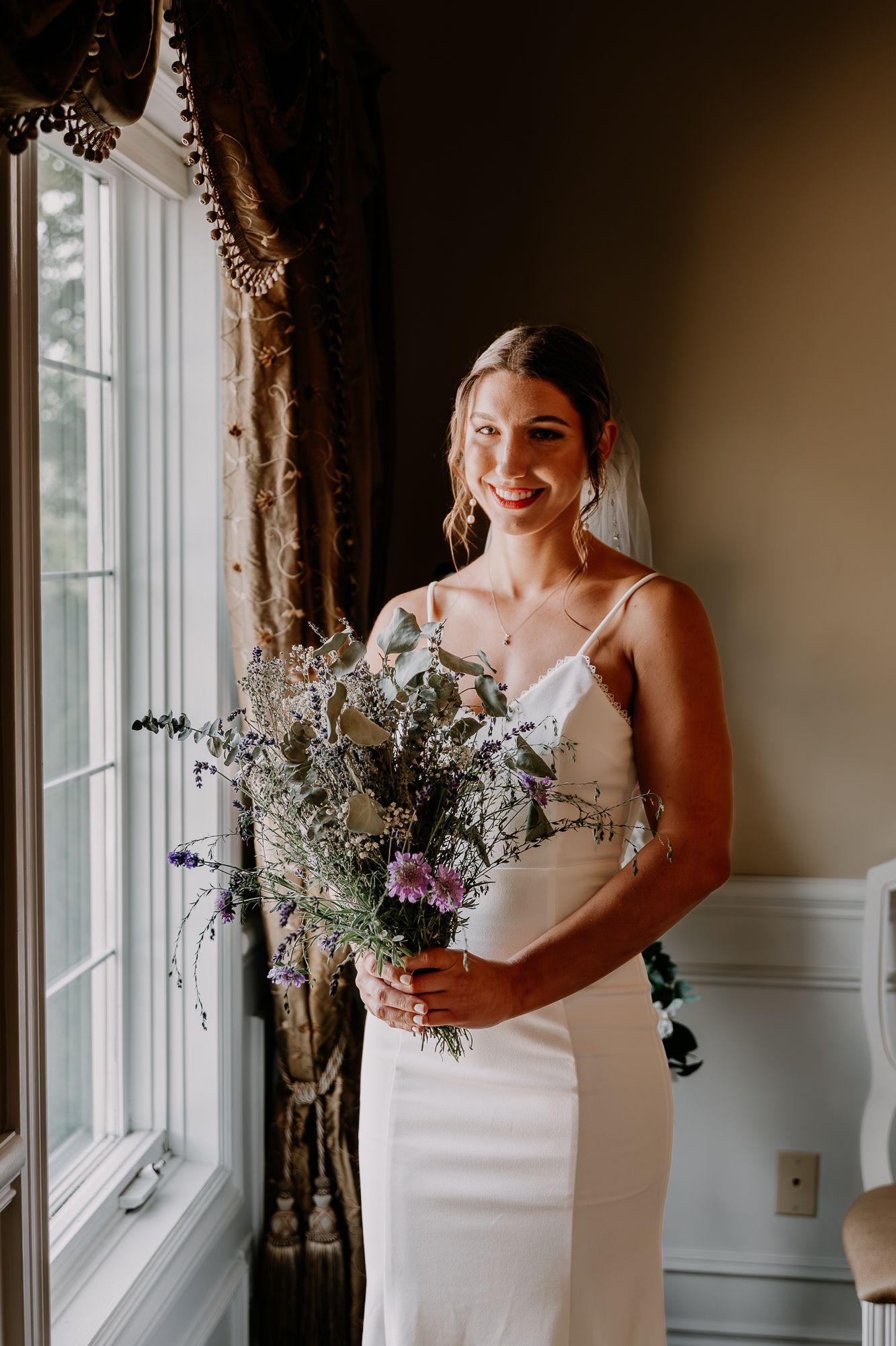 Bride in elegant white dress holds wildflower bouquet while standing by window.