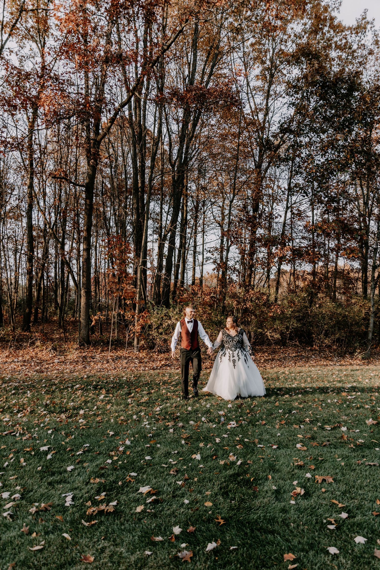 Wedding couple holds hands in autumn forest clearing surrounded by fall foliage.