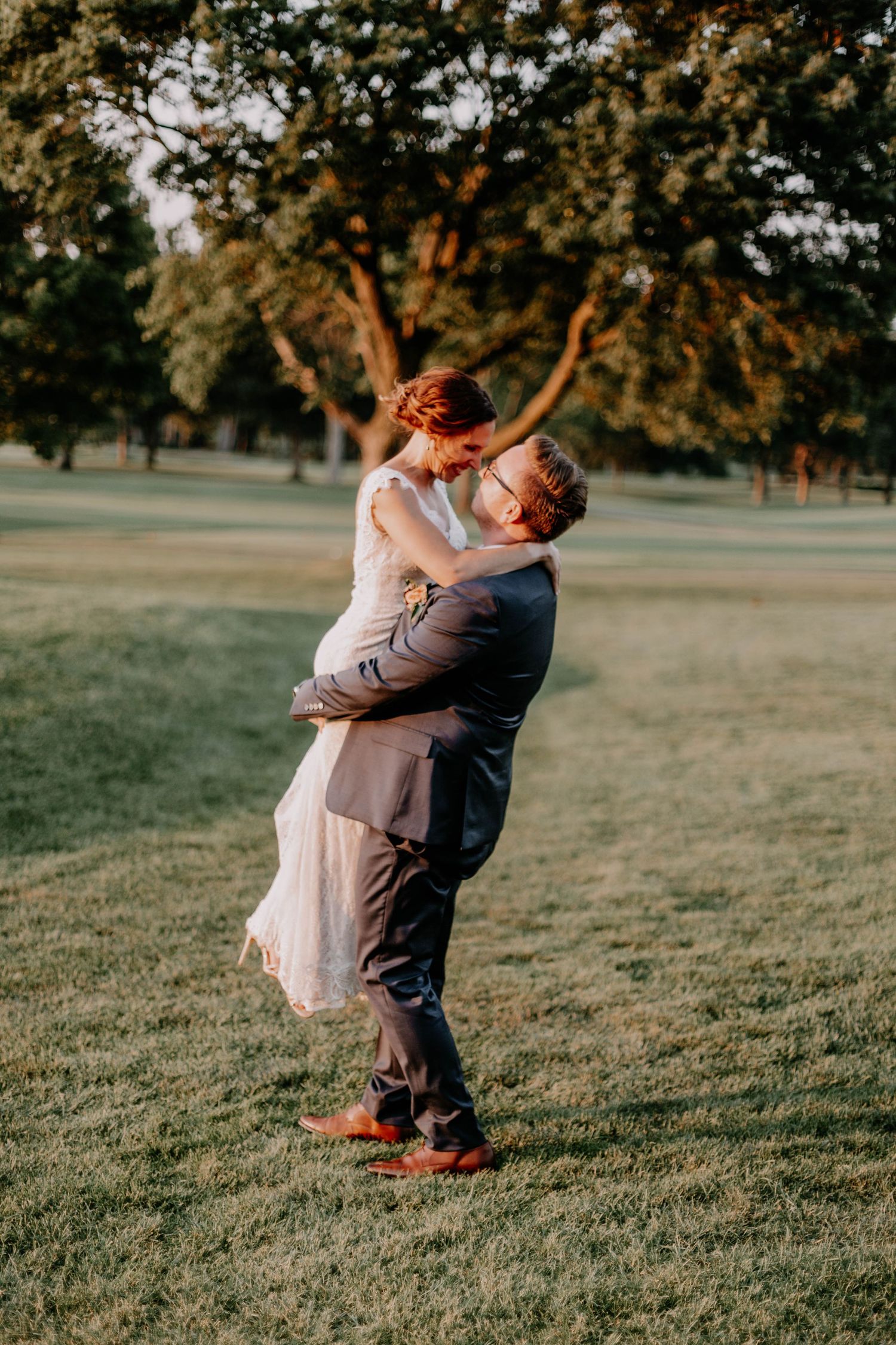 A groom lifts his bride in a romantic embrace during their sunset wedding photoshoot in a grassy field.