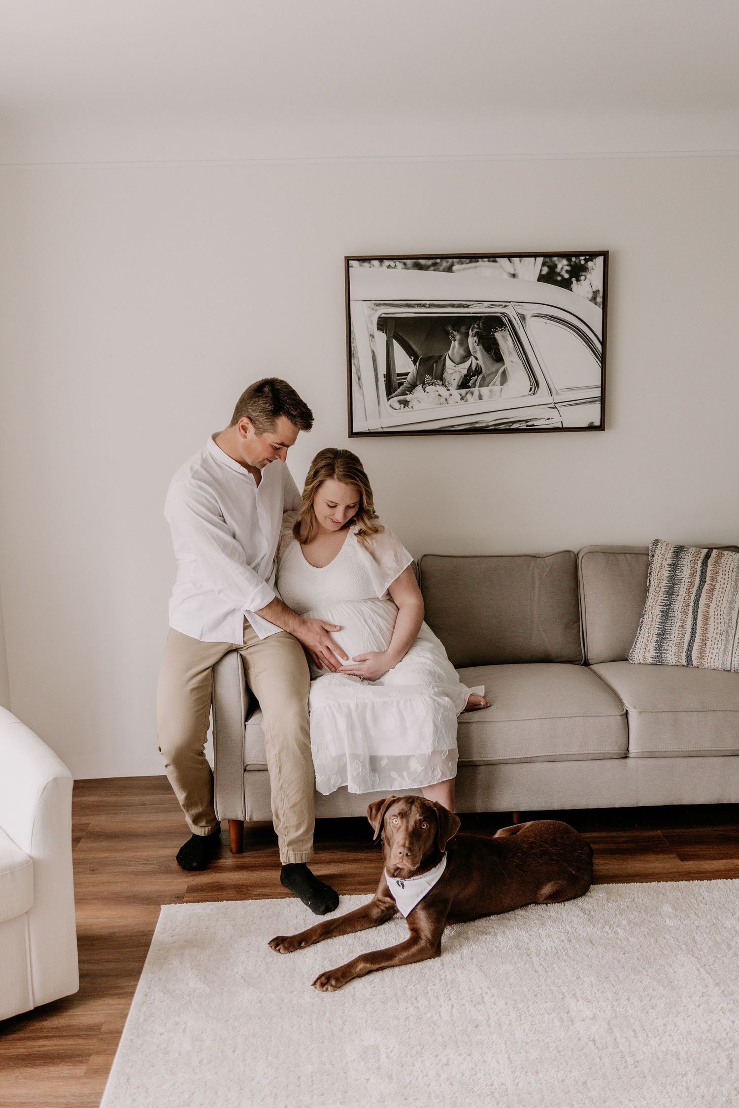 Couple tenderly touching pregnant belly seated on gray sofa with chocolate Labrador dog lying on white rug in cozy living room.