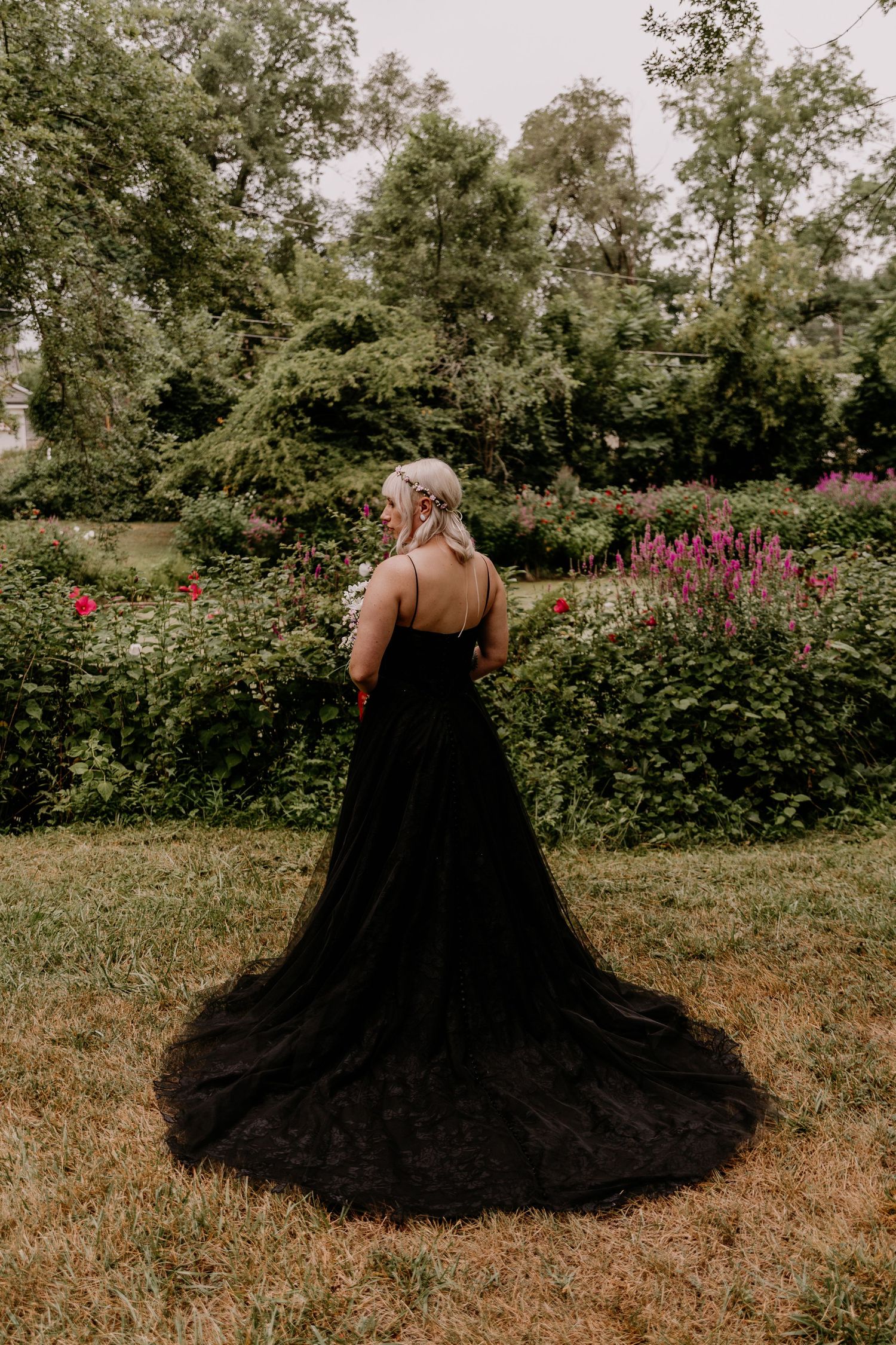 Woman in elegant black tulle gown with train stands in lush garden, viewed from behind.