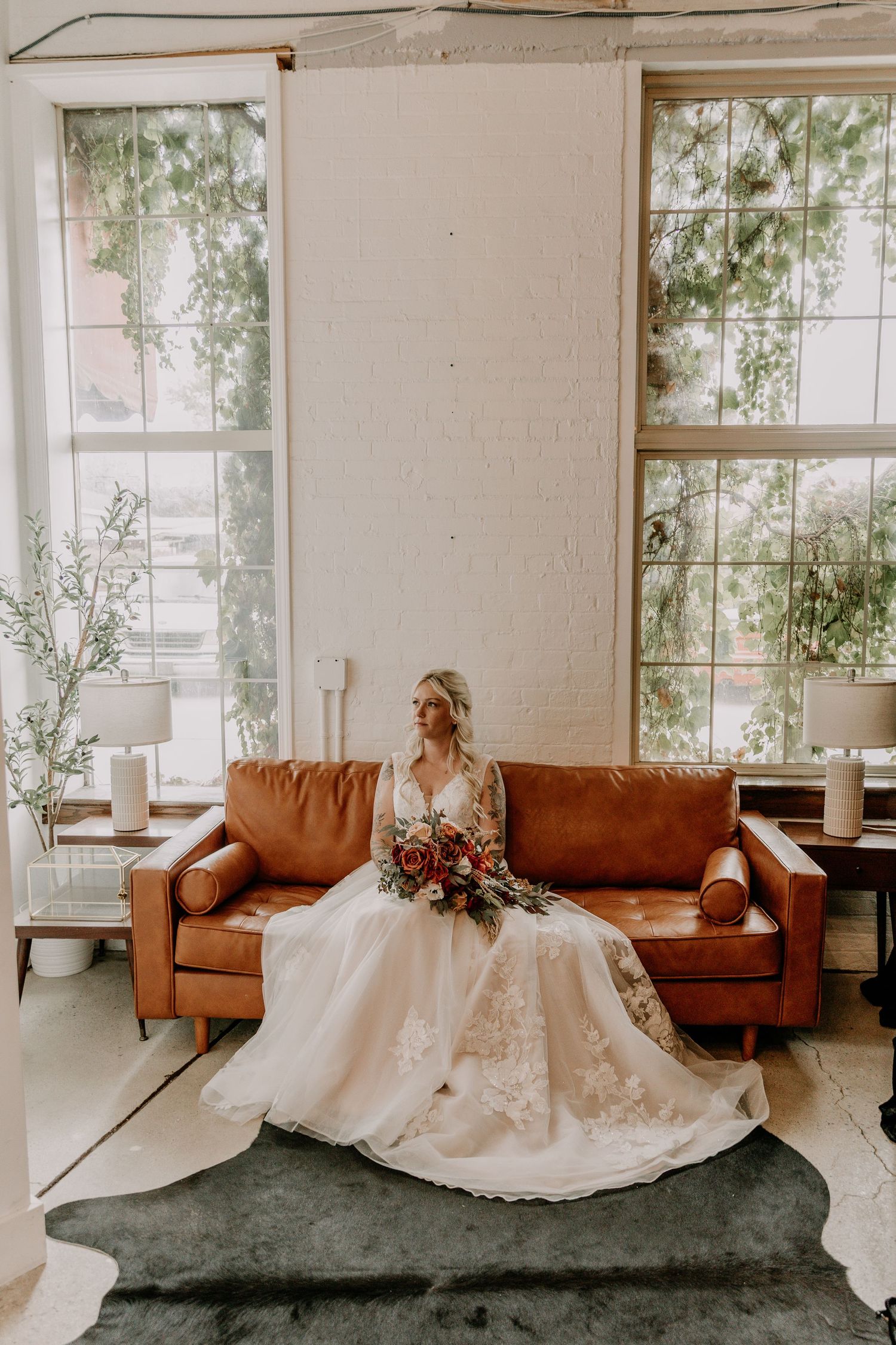 A bride in a flowing white wedding gown sits gracefully on a brown leather couch near bright sunlit windows.