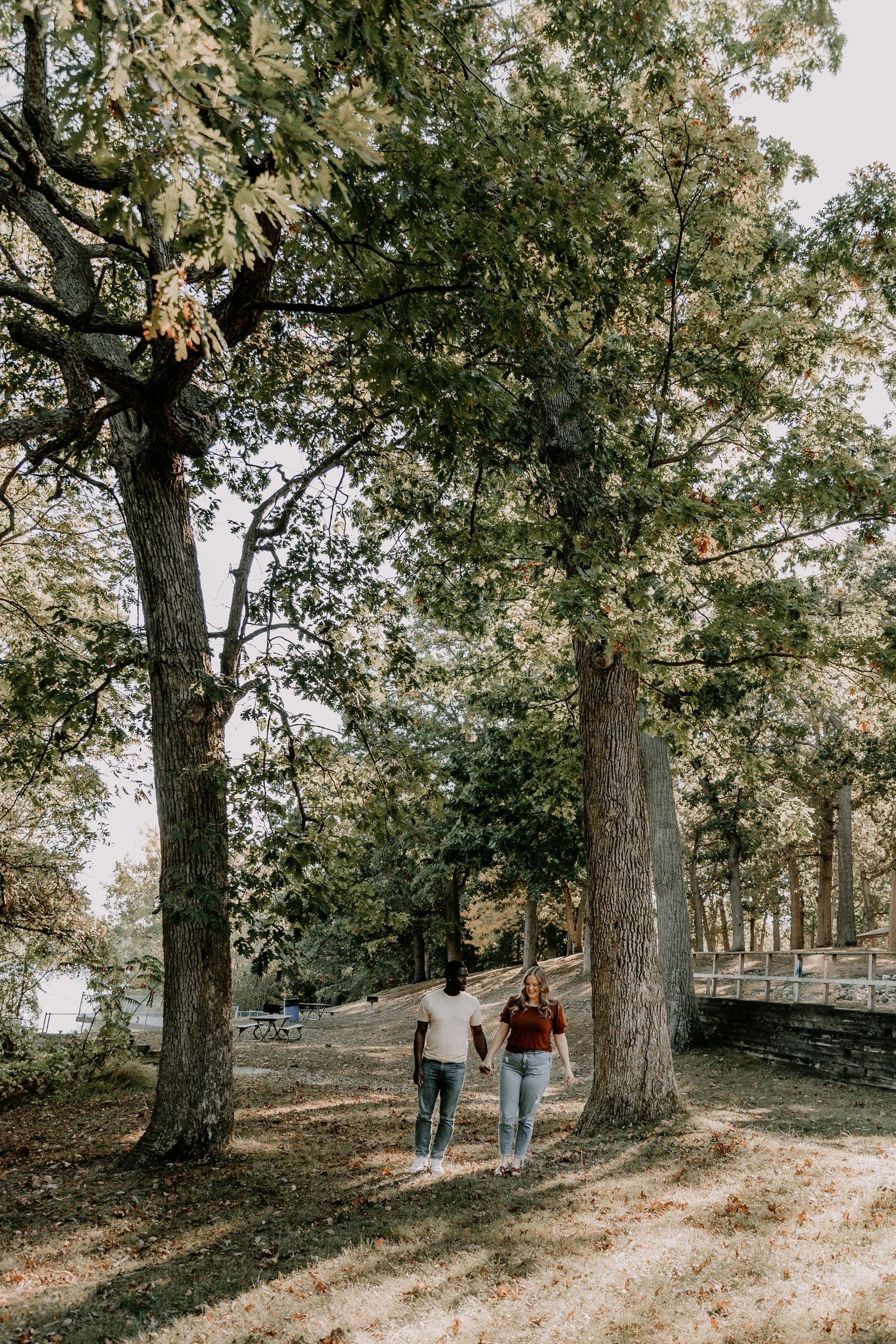 A couple walks along a tree-lined path in a scenic park setting during autumn with dappled sunlight filtering through leaves.