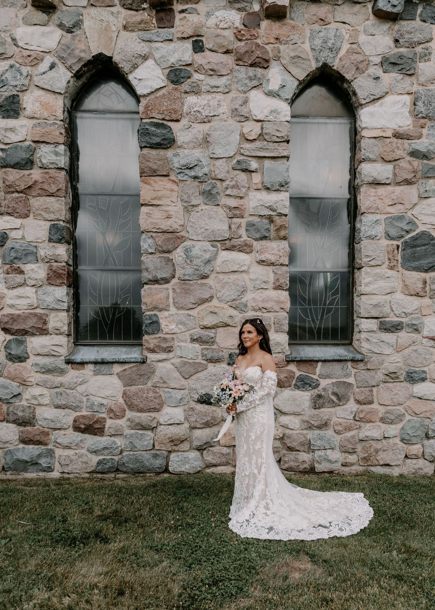 A figure in a lace wedding dress stands against a rustic stone church wall with two gothic arched windows.