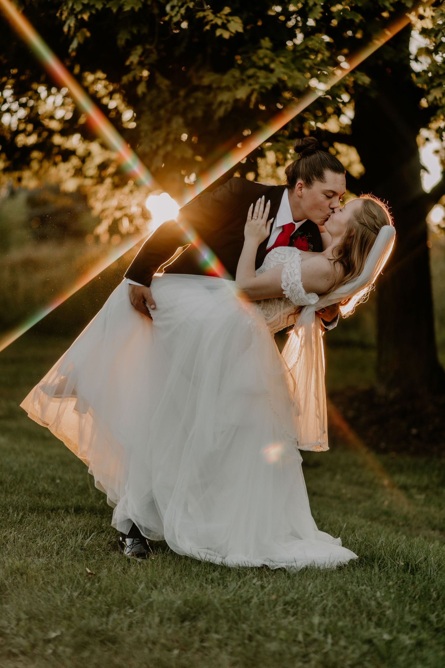Groom dips bride for a kiss at golden hour sunset, with light flares framing the romantic wedding moment outdoors.