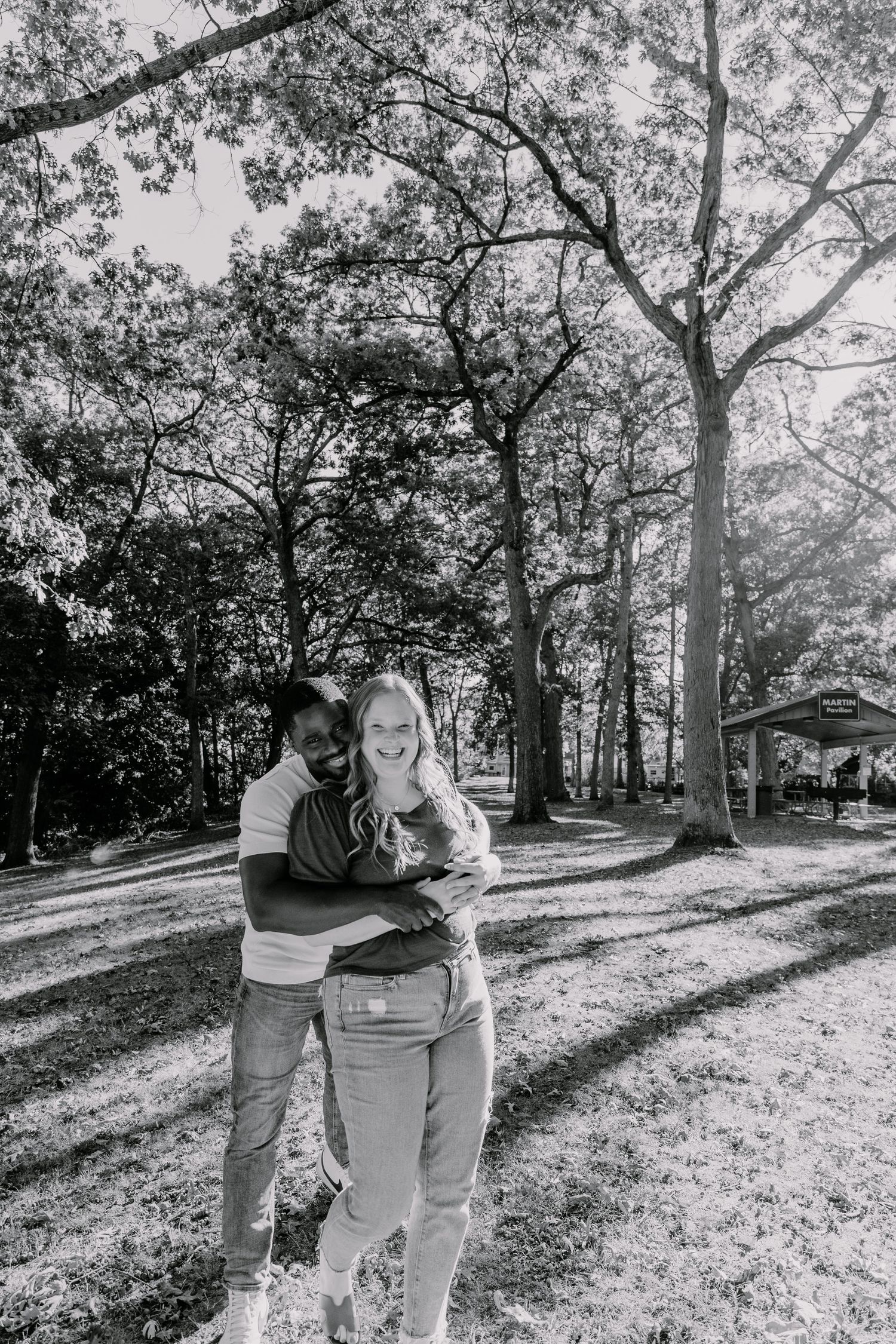 A couple walks together through a sun-dappled park on an autumn day, captured in black and white photography.