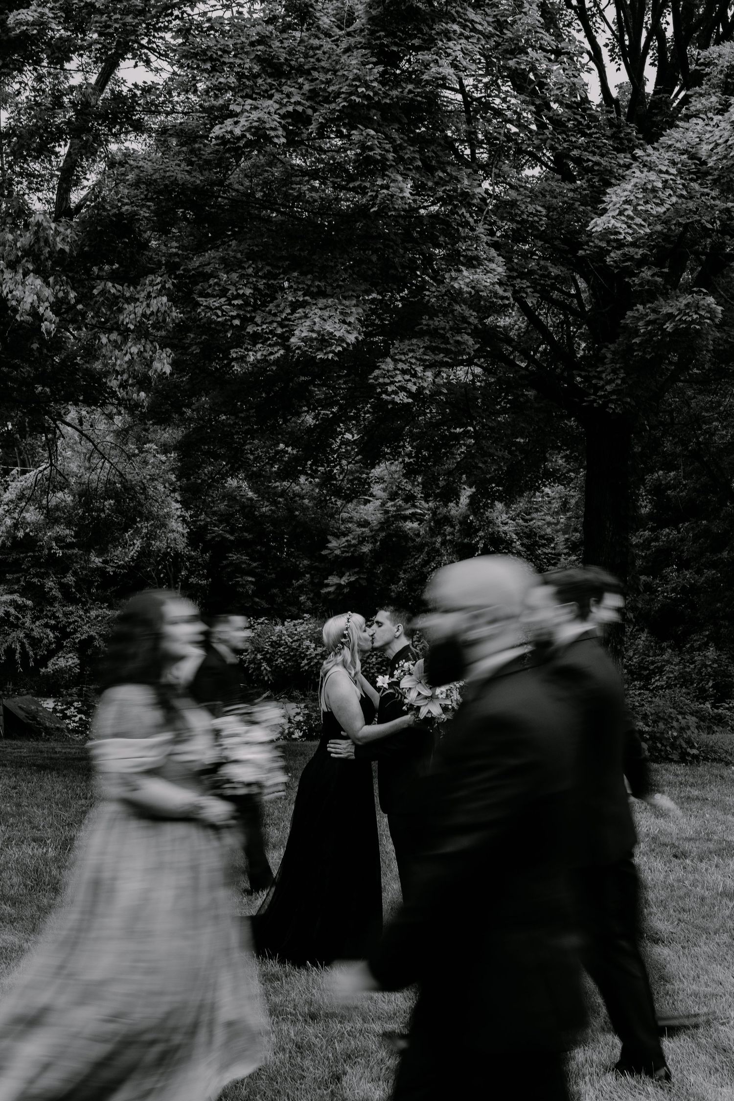 Black and white wedding photo with blurred guests in foreground as couple kisses surrounded by lush trees.