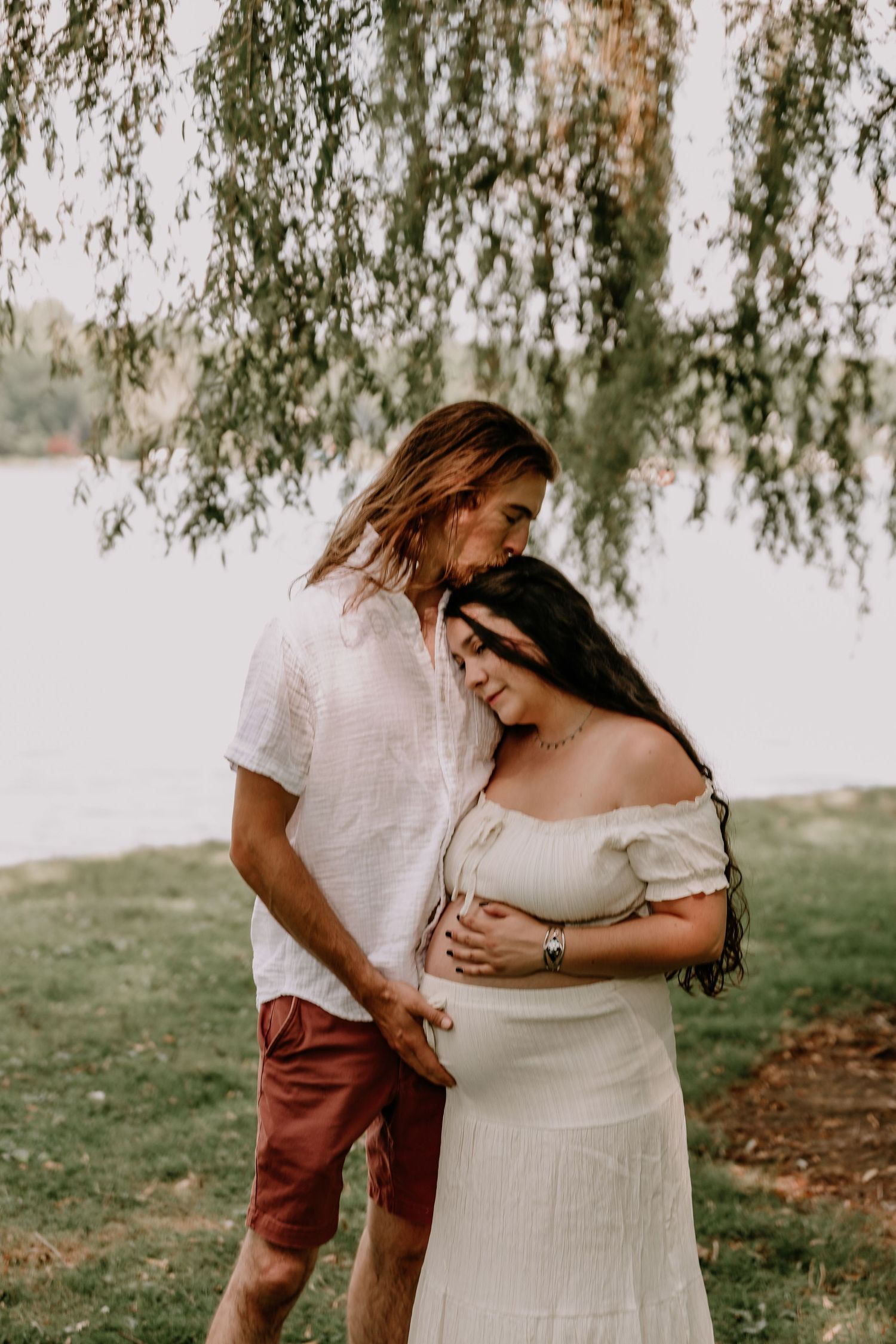 Couple sharing a tender maternity moment under a weeping willow tree by a lake, woman in white dress.