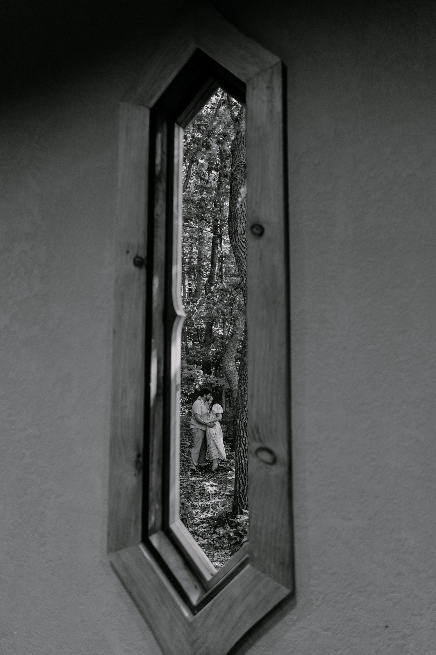 Black and white photo of a couple embracing in a forest, viewed through a narrow arched wooden window.