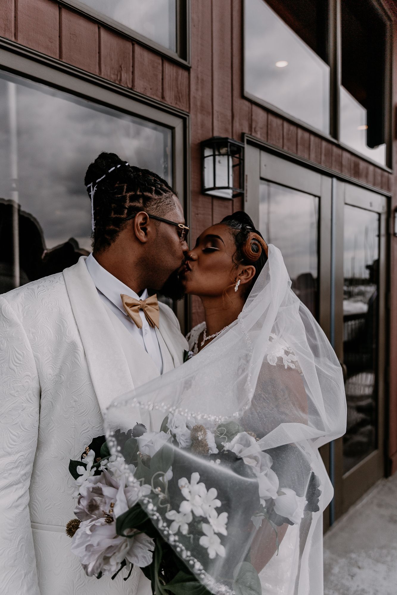 Romantic wedding couple sharing a tender kiss outside a rustic brown building.