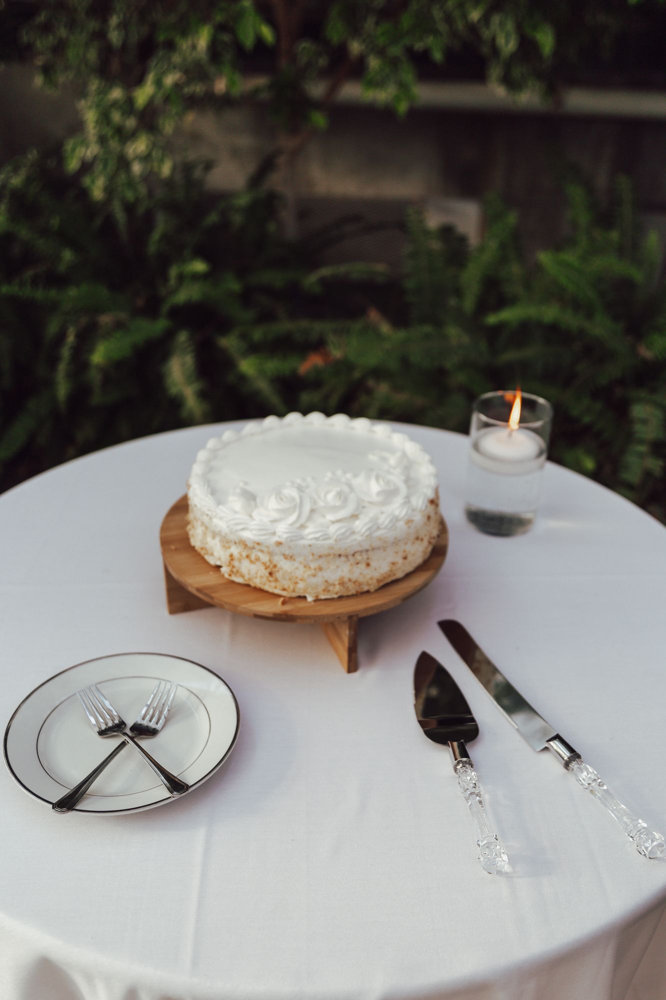 A white wedding cake with whipped cream frosting sits on a wooden stand with elegant serving utensils and candle nearby.