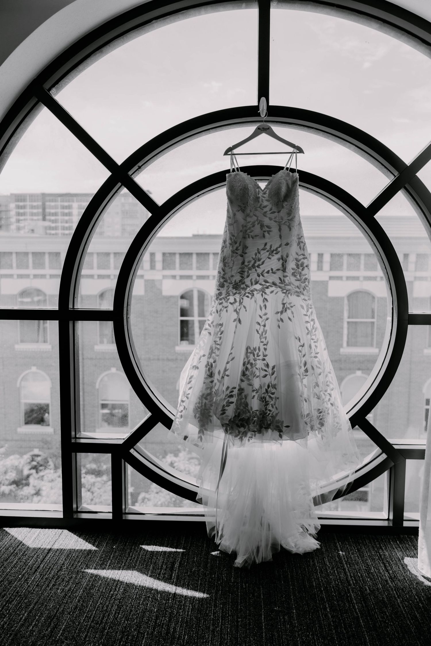 Wedding dress hanging in front of an elegant arched window in black and white photography.