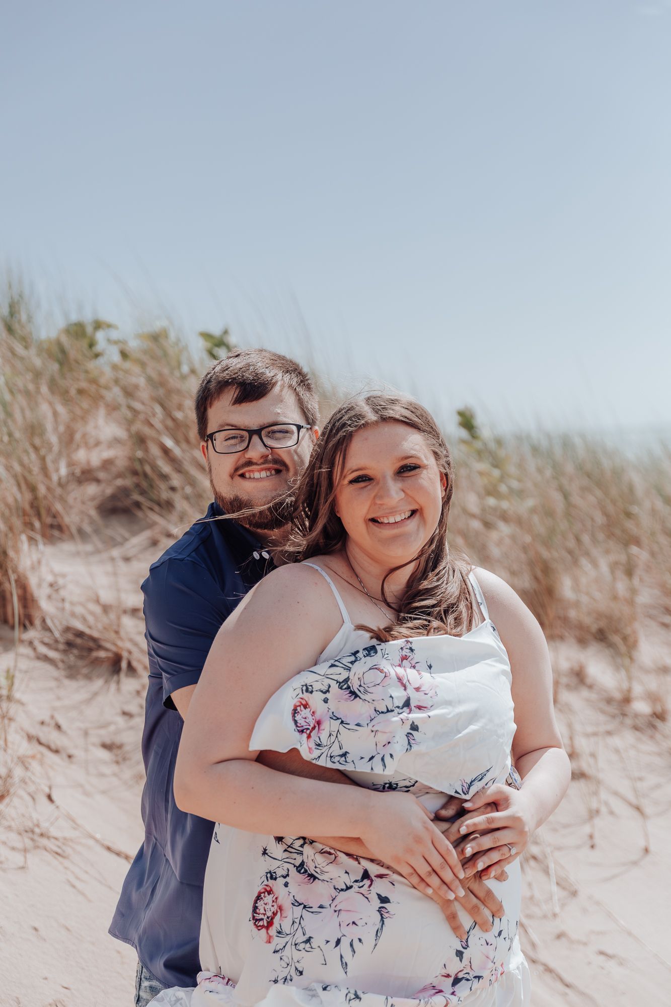 A couple shares a tender moment while standing on sandy beach dunes on a bright sunny day.