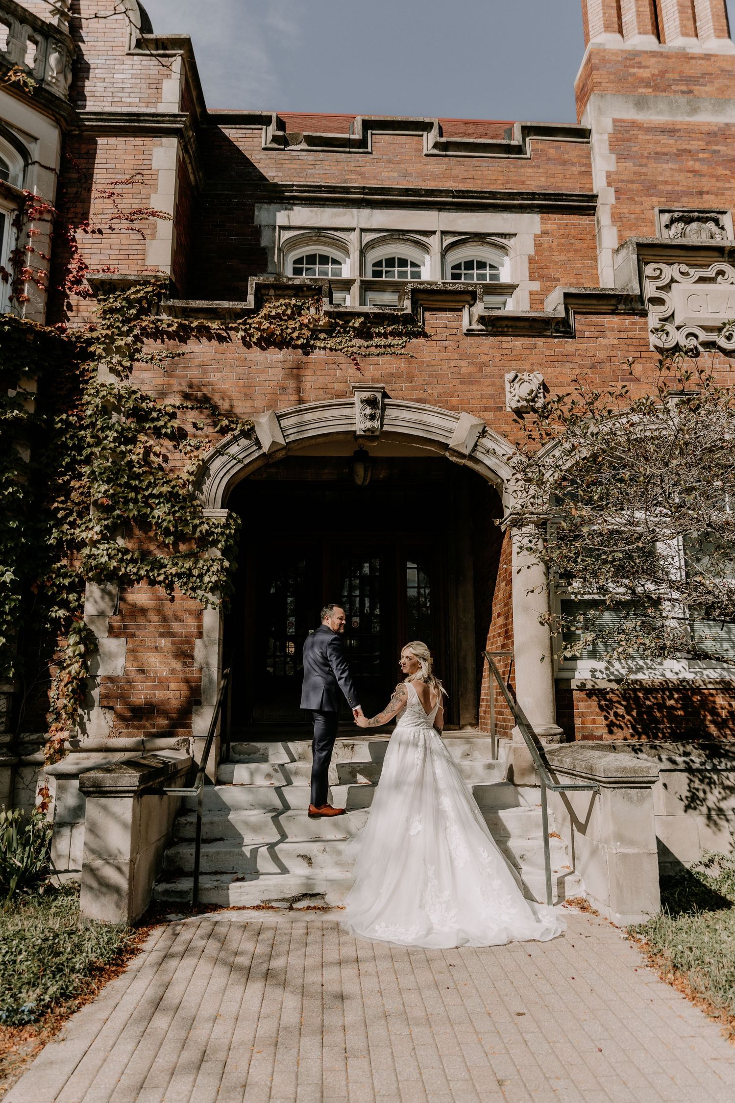A couple in formal attire walking together outside a historic brick building with arched doorways.