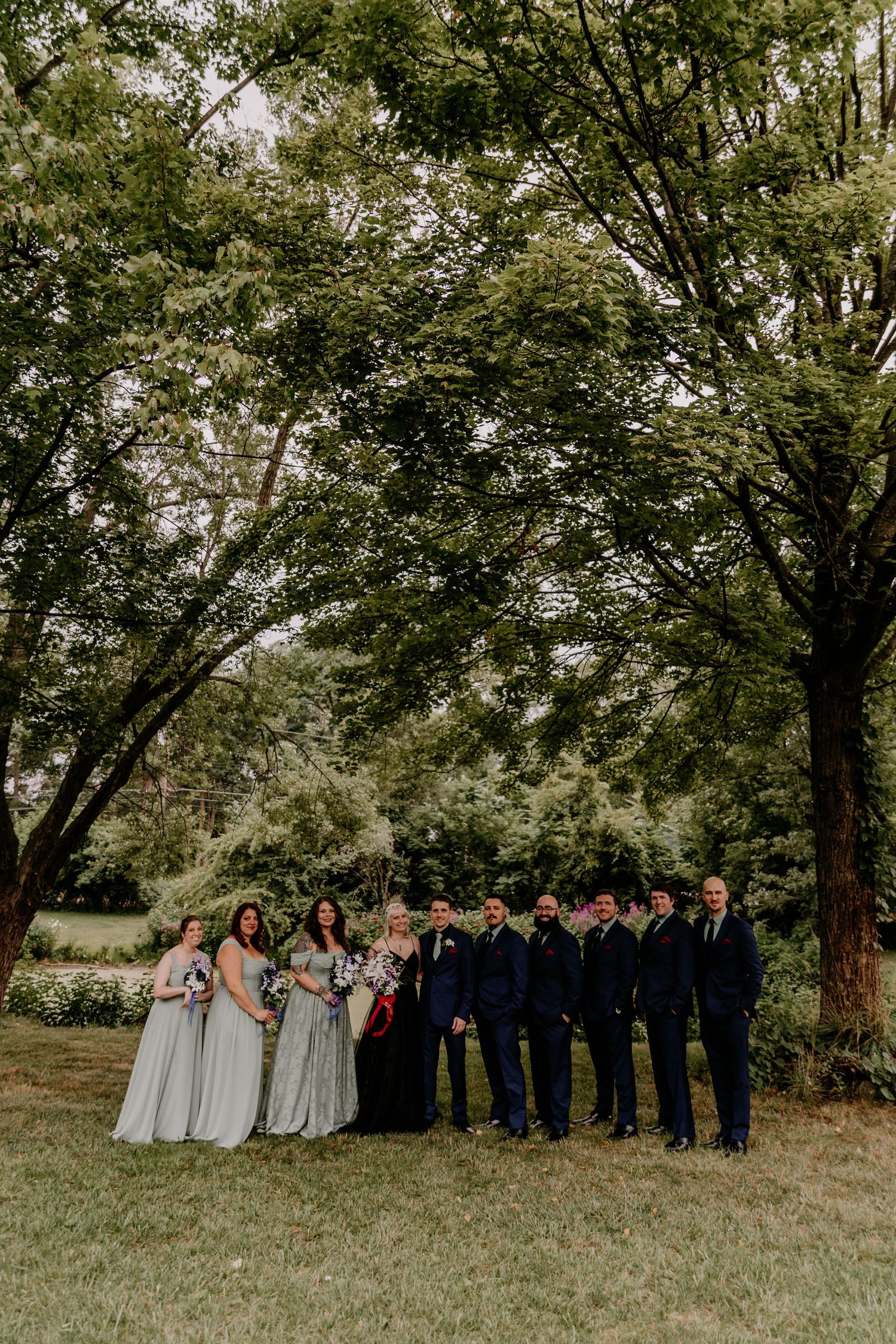 Wedding party poses outdoors under lush green trees, bridesmaids in sage gowns and groomsmen in navy suits flanking the couple.
