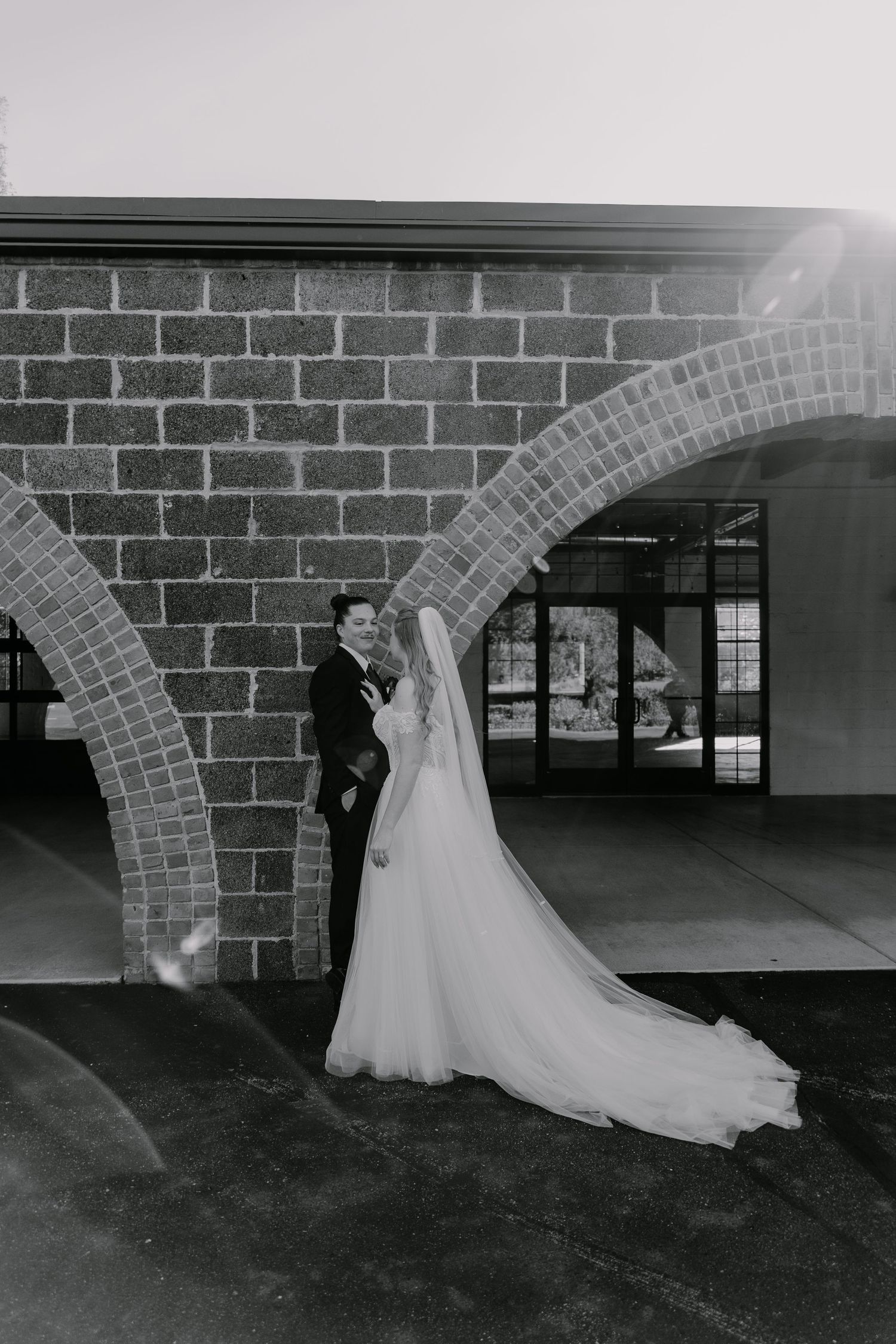 Black and white photo of a bride and groom posing under a brick arch, bride's veil flowing gracefully.