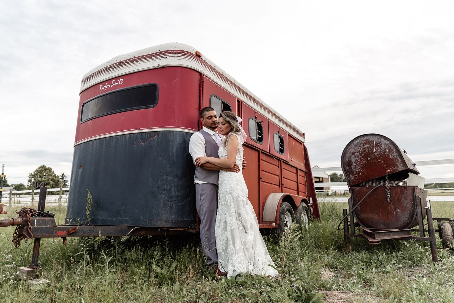 Newlyweds embracing in front of vintage red horse trailer in rustic field setting.